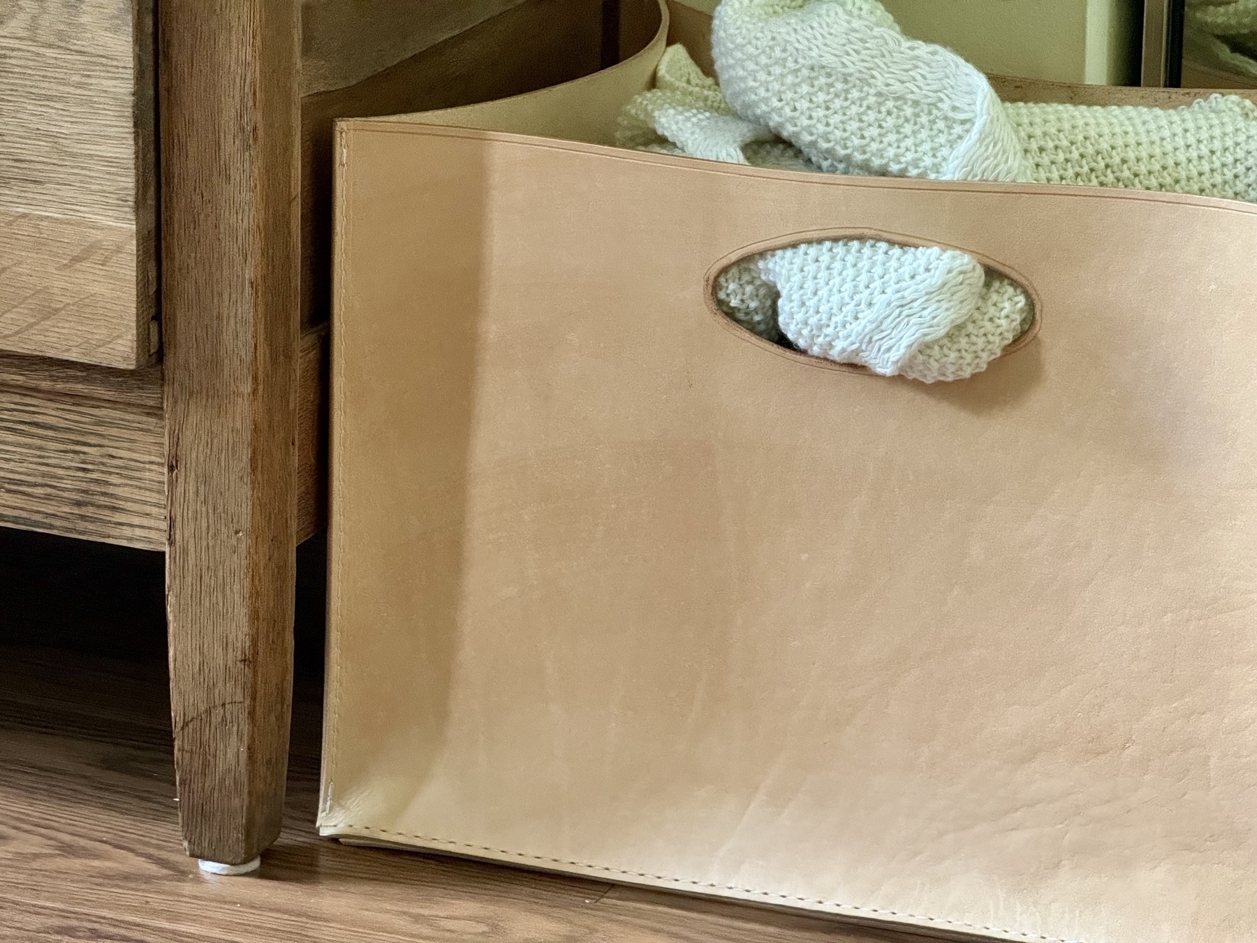 A beige rectangular leather storage basket with a knitted item inside is placed next to a wooden piece of furniture on a wooden floor.