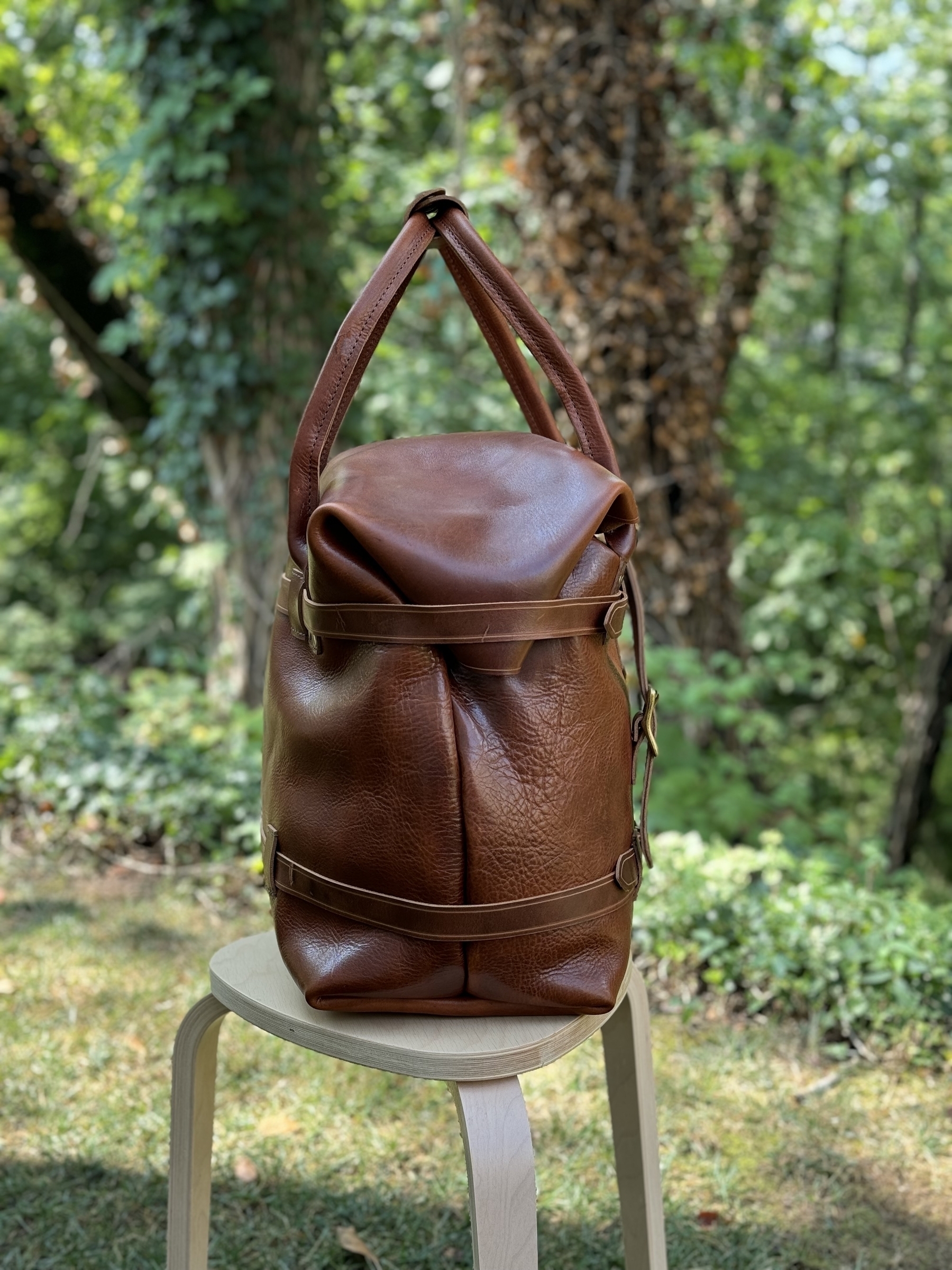 A brown leather weekend bag bag is sitting on a wooden stool outdoors with a forested background.