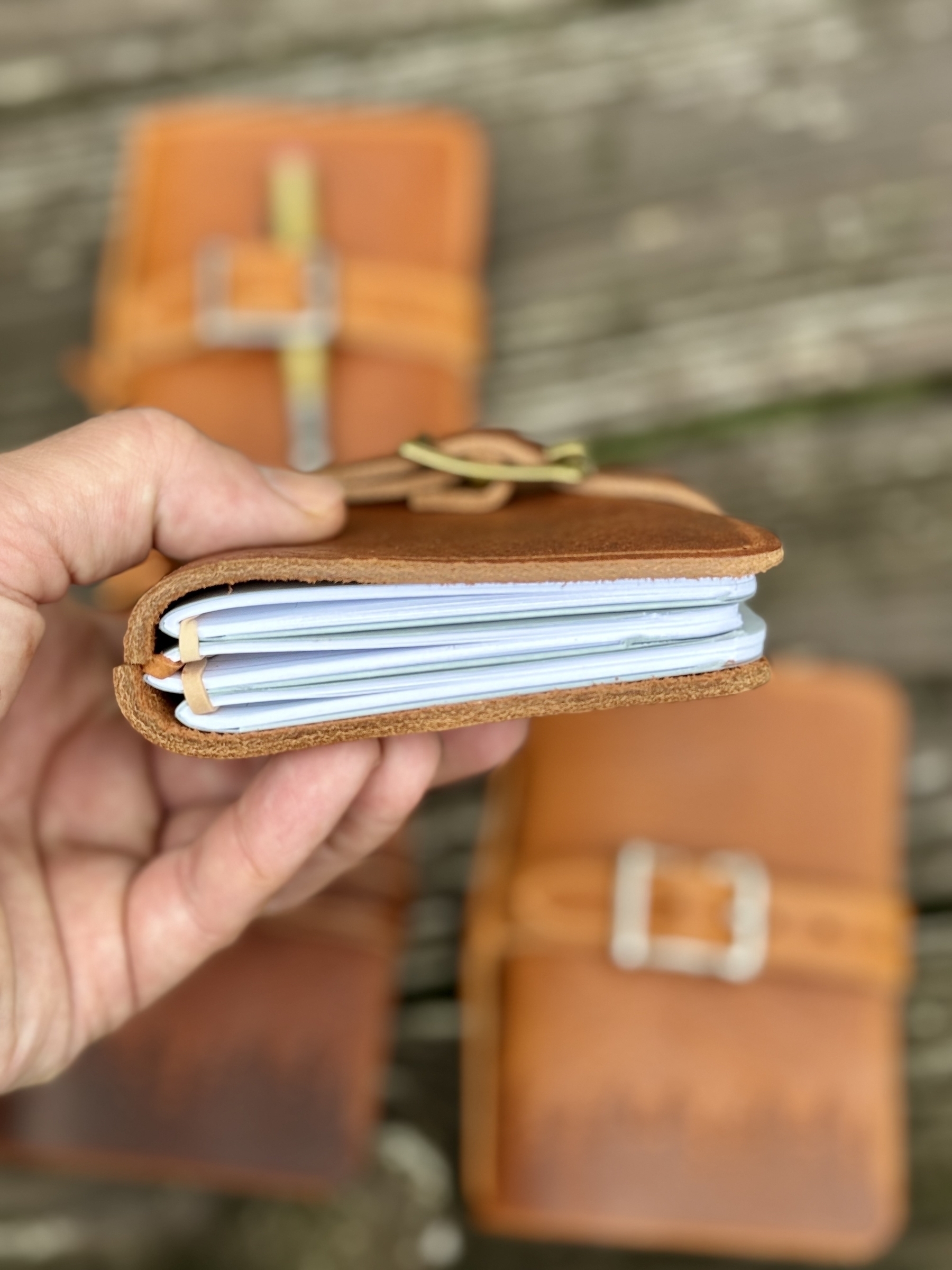 A hand is holding a small leather-bound notebook with several pages, and two similar notebooks are placed on a wooden surface in the background.