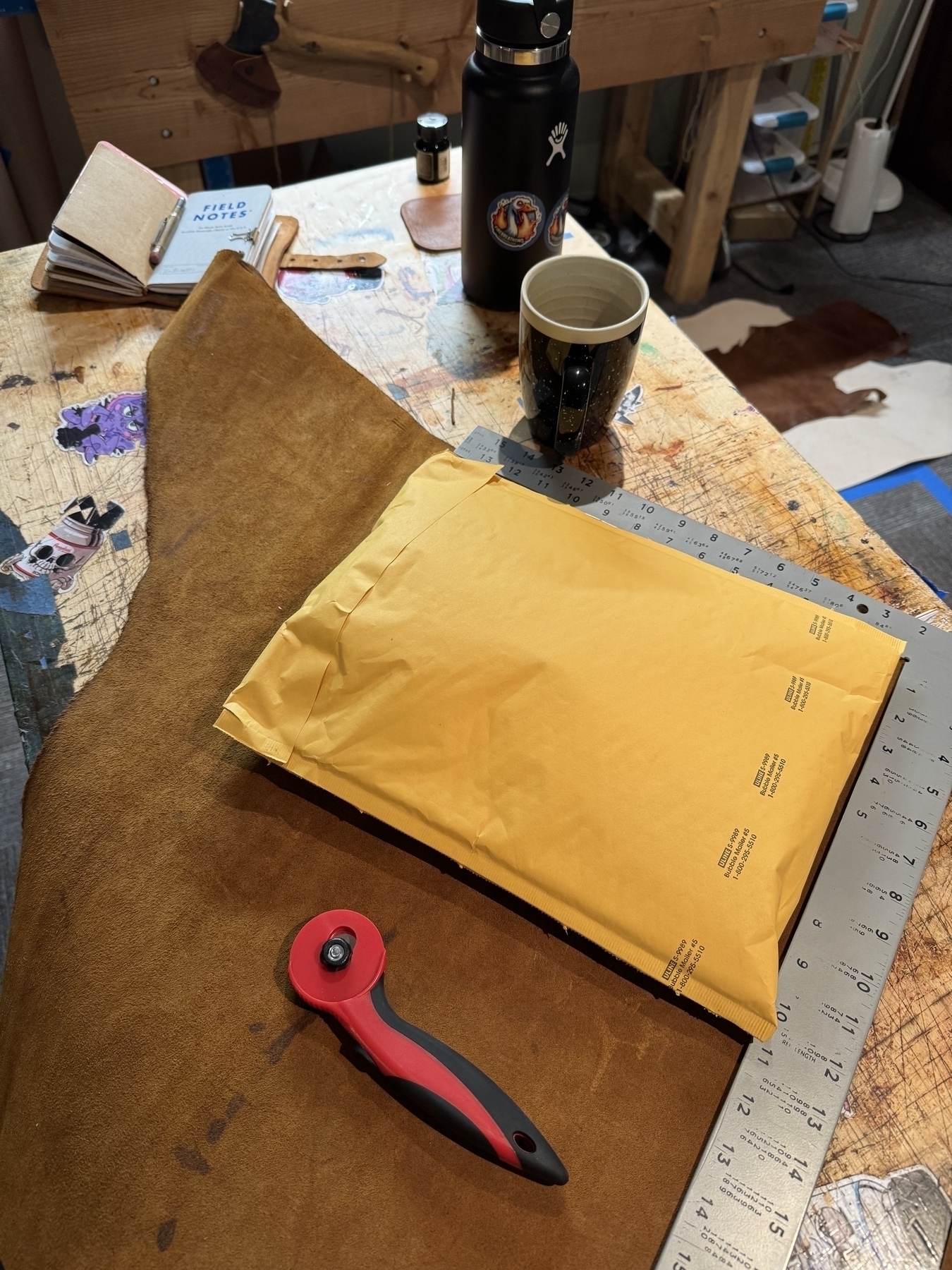 A workbench with a cutting tool, measuring ruler, yellow envelope, and a mug is set up for crafting or mailing tasks.