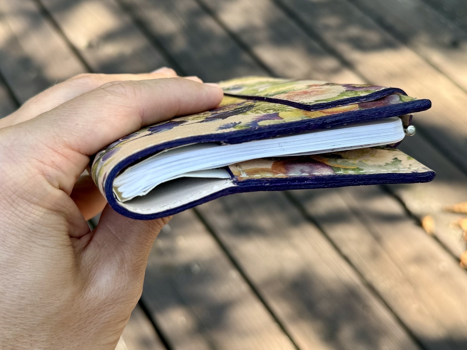 A hand is holding a small, colorful floral-patterned notebook or wallet with a curvy edge.