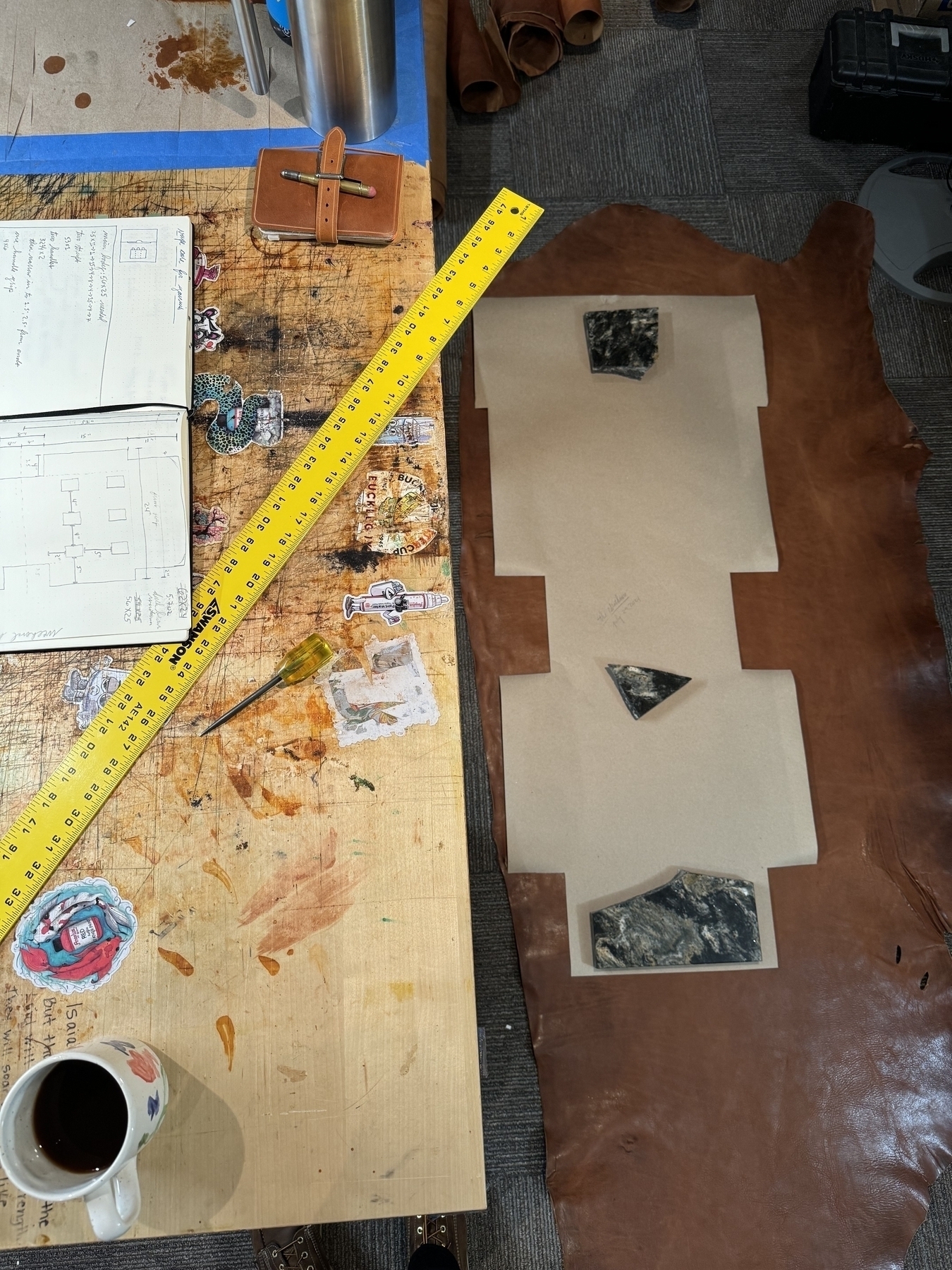 A workspace featuring a long piece of leather, a yellow measuring tape, an open notebook with sketches, a coffee cup, and various tools and materials scattered around.