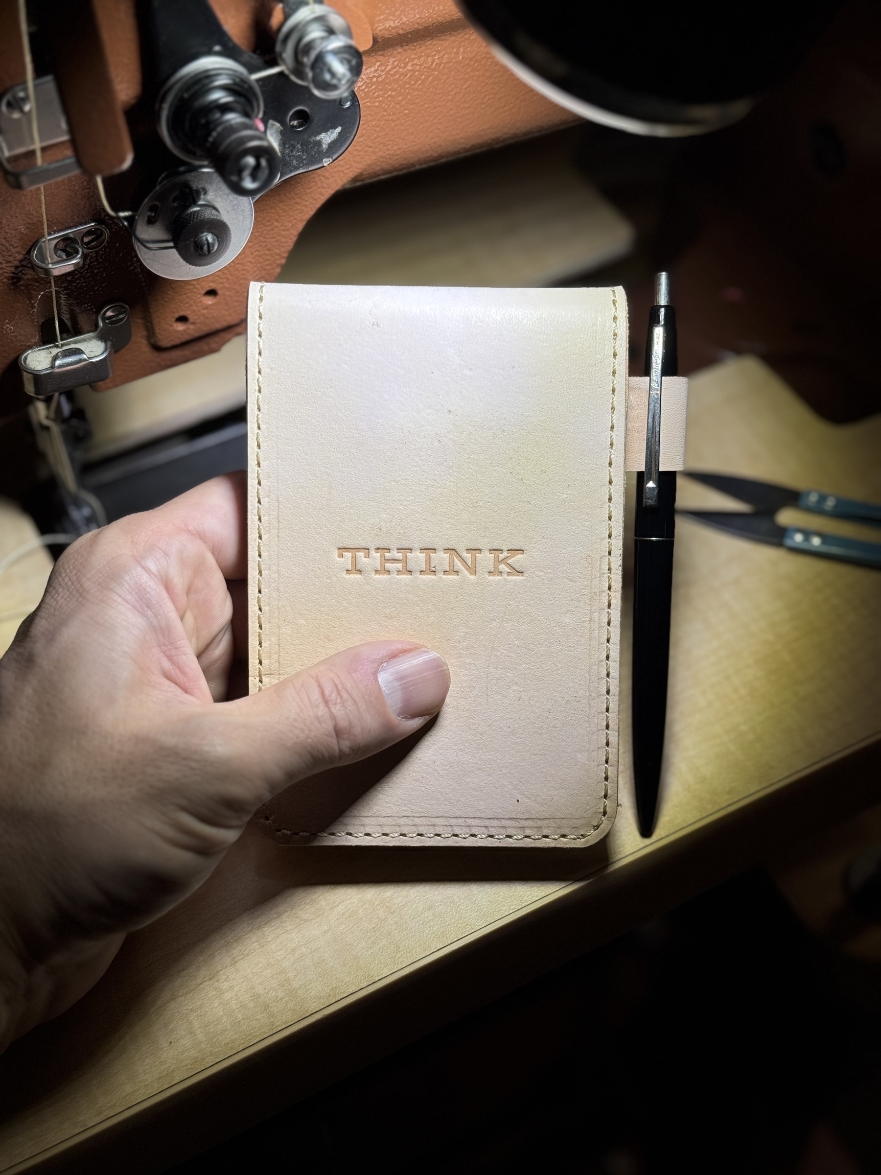 A hand is holding a small leather notebook embossed with the word "THINK," next to a black pen on a table with sewing equipment in the background.