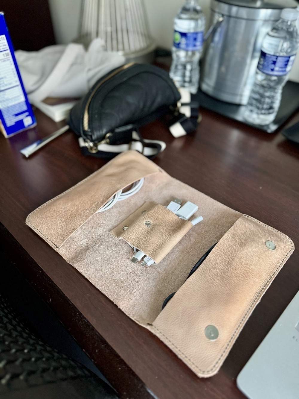 A leather organizer on a desk holds various cables, with water bottles, a bag, and other items in the background.