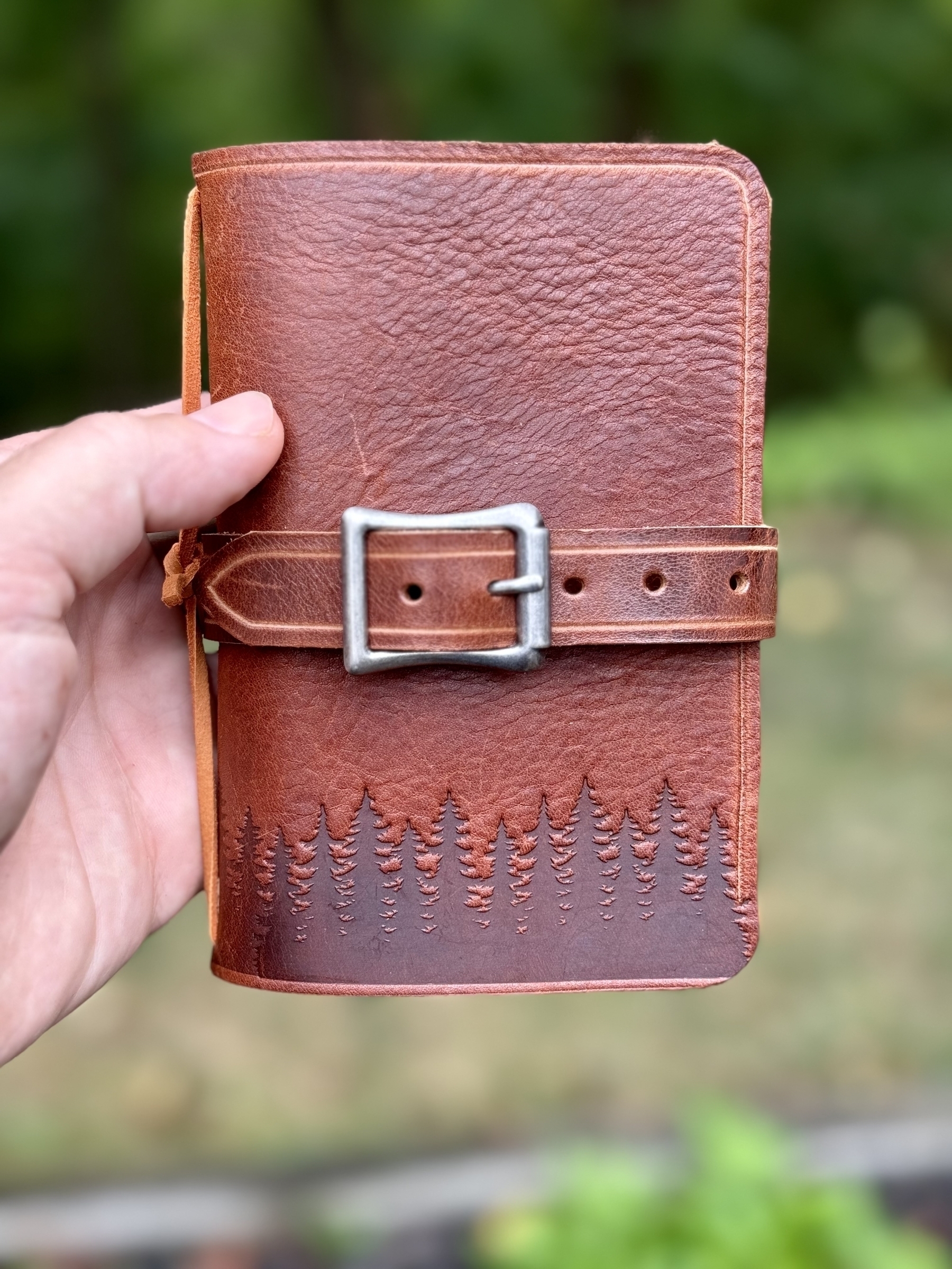 A hand is holding a brown leather notebook with an embossed tree design and a metal buckle closure.