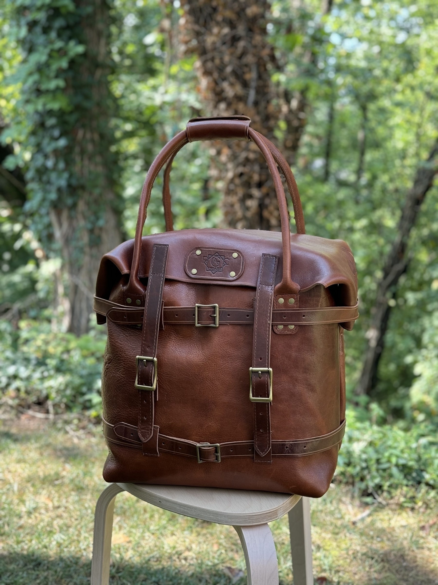 A brown leather weekend bag with buckles is placed on a wooden stool outdoors in a lush, green environment.