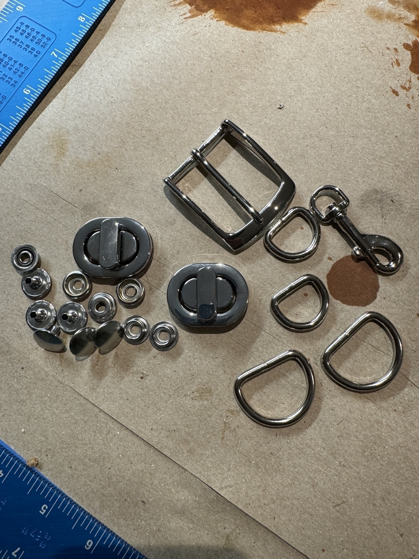 The image shows various metal hardware pieces, including buckles, D-rings, and snap fasteners, arranged on a beige surface next to blue measuring tools.