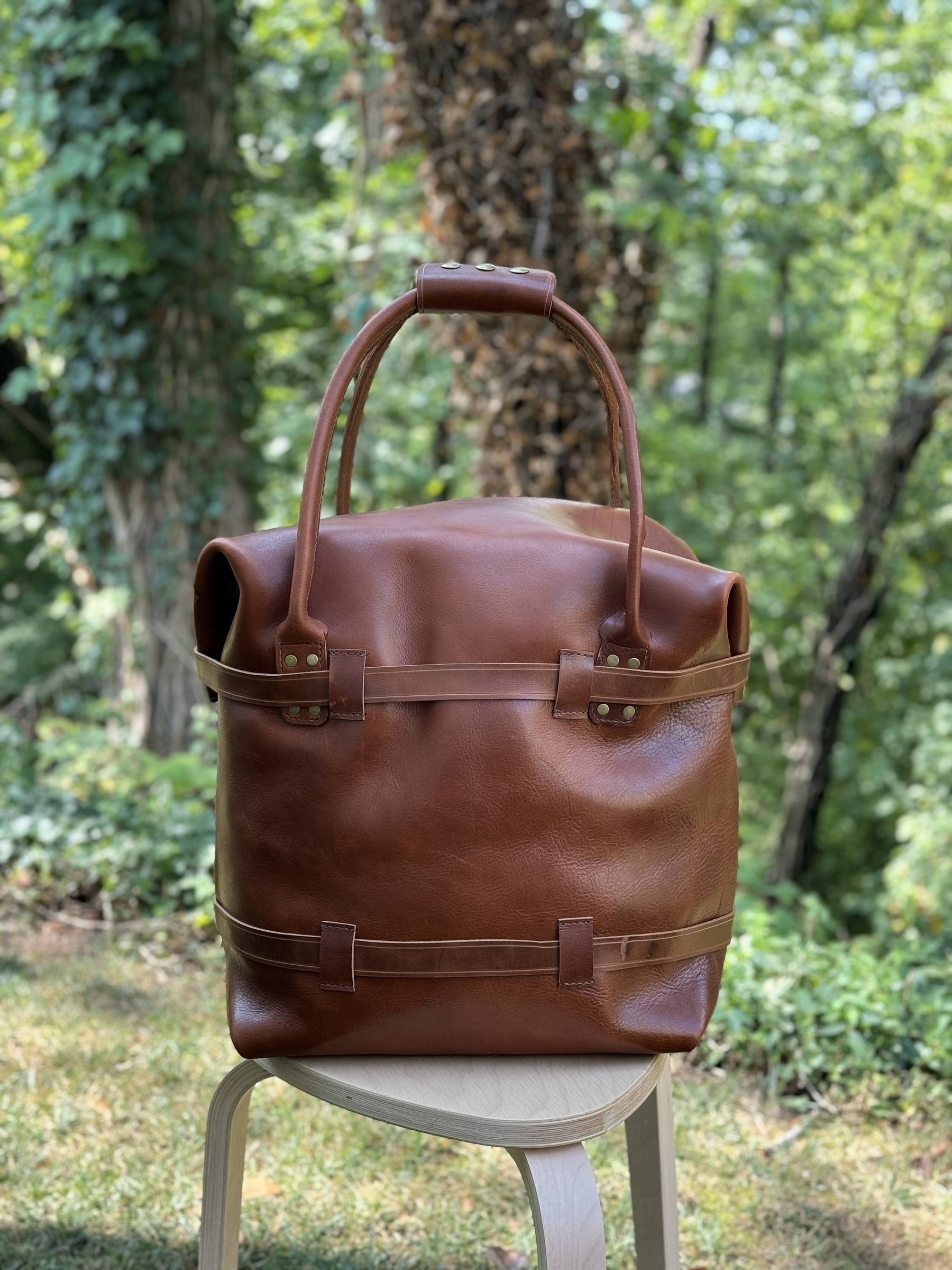 A large brown leather weekend bag with rolled handles is placed on a wooden stool in an outdoor setting with greenery in the background.