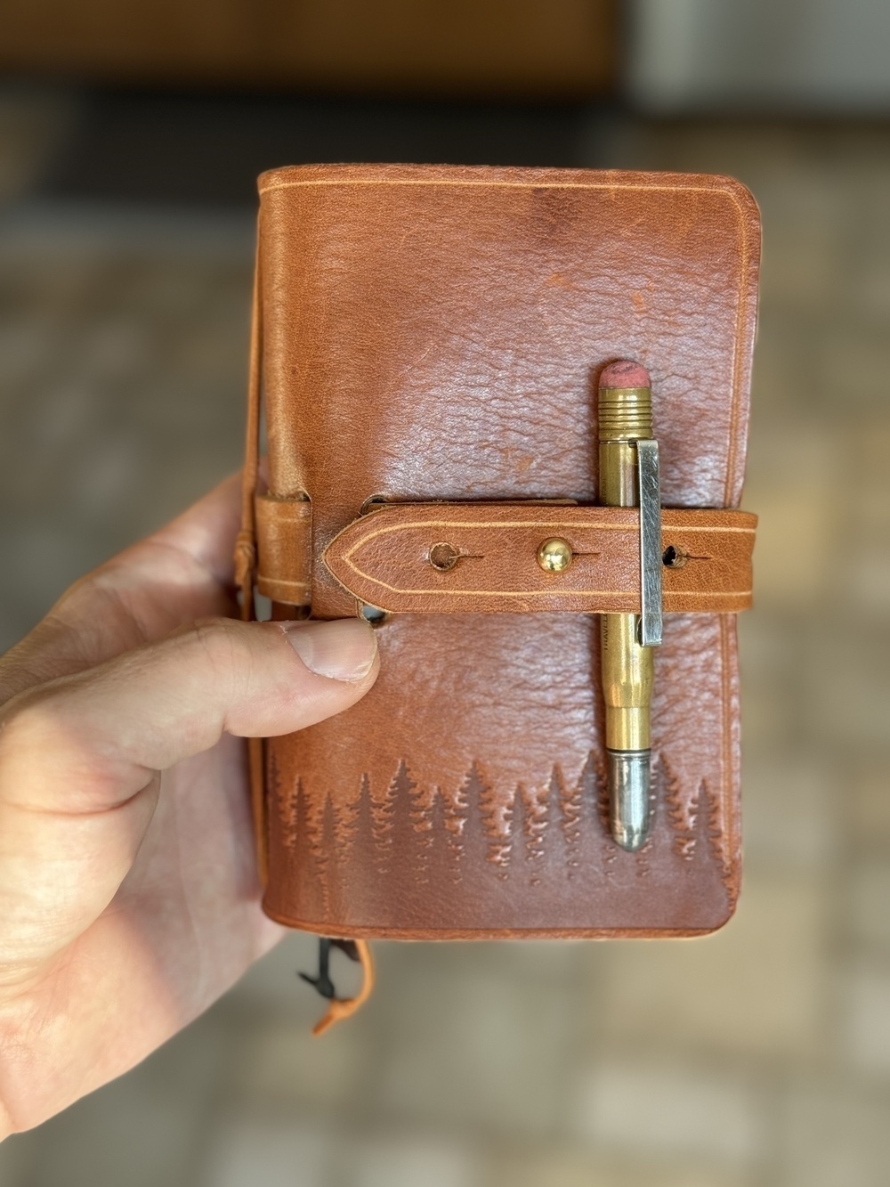 A hand is holding a brown leather-bound notebook with tree engravings, secured with a strap holding a pencil.