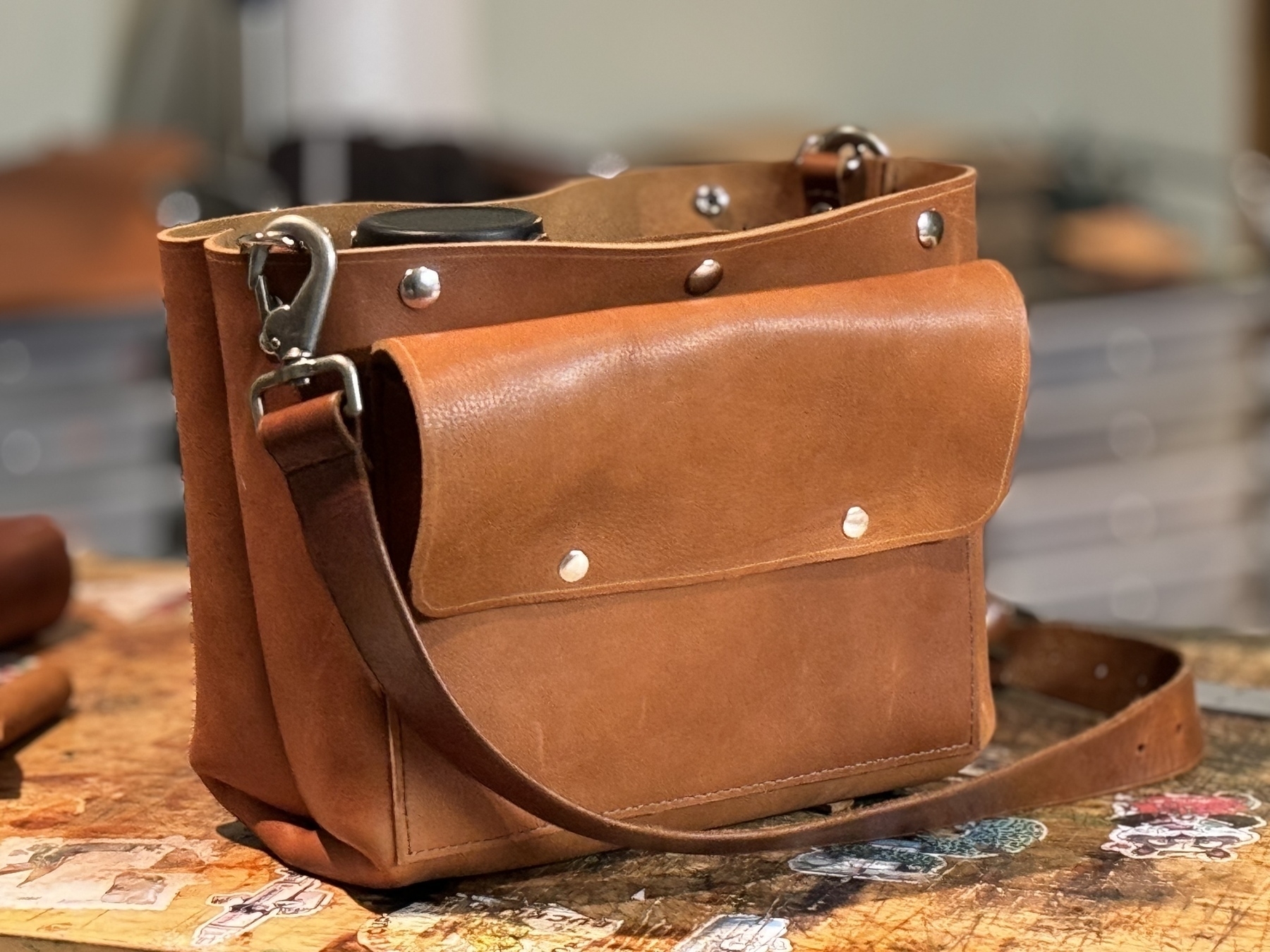 A brown leather shoulder bag with metal accents and a front pocket is placed on a wooden surface.