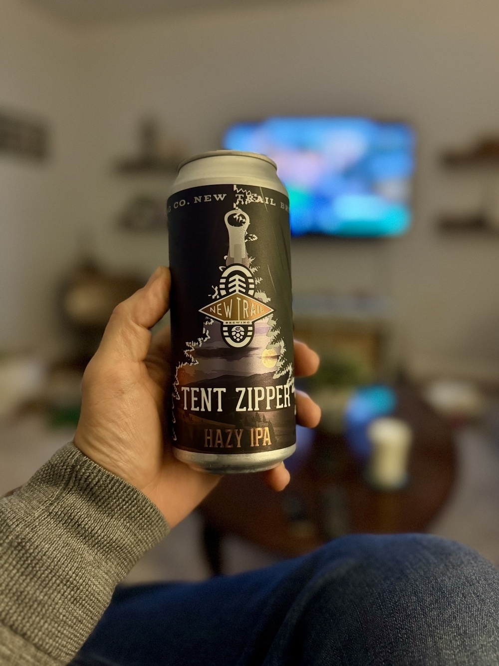 A person is holding a can of "Tent Zipper Hazy IPA" beer in a living room with a TV in the background.