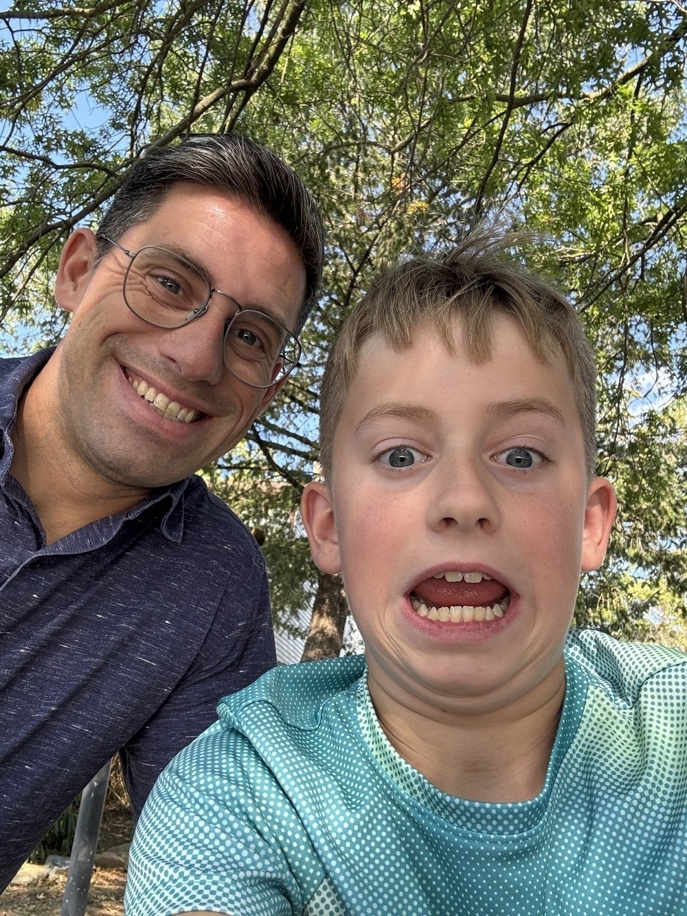 A man and a child are taking a playful selfie under a tree.