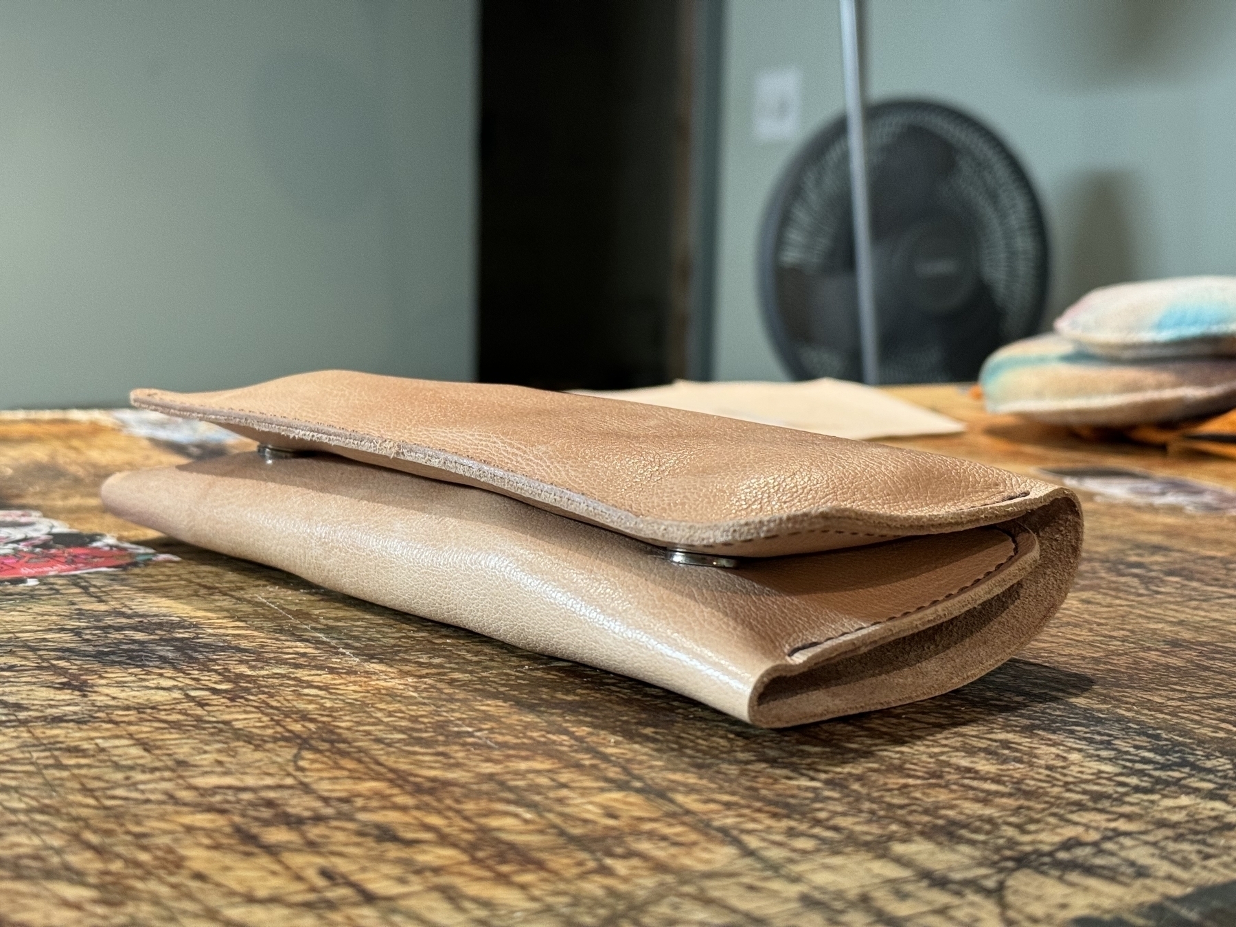 A folded brown leather wallet is resting on a wooden surface with a fan and other blurred items in the background.