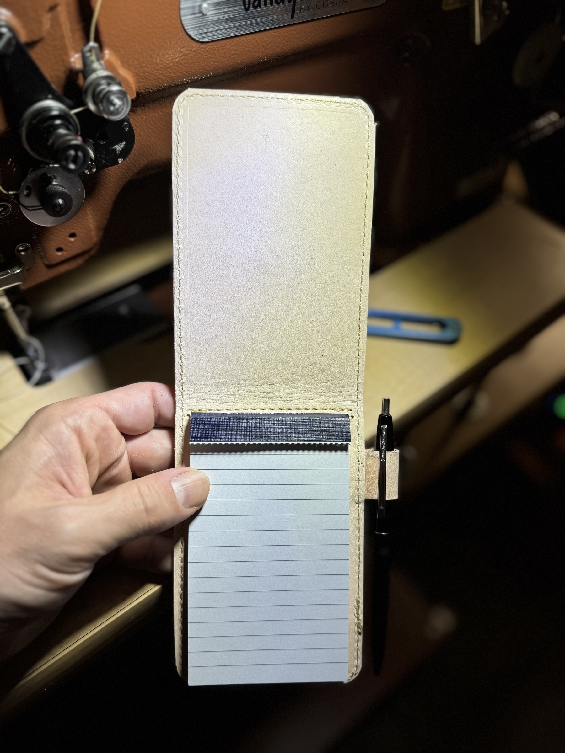 A hand is holding an open leather notepad with lined paper, featuring a pen attached to its cover.