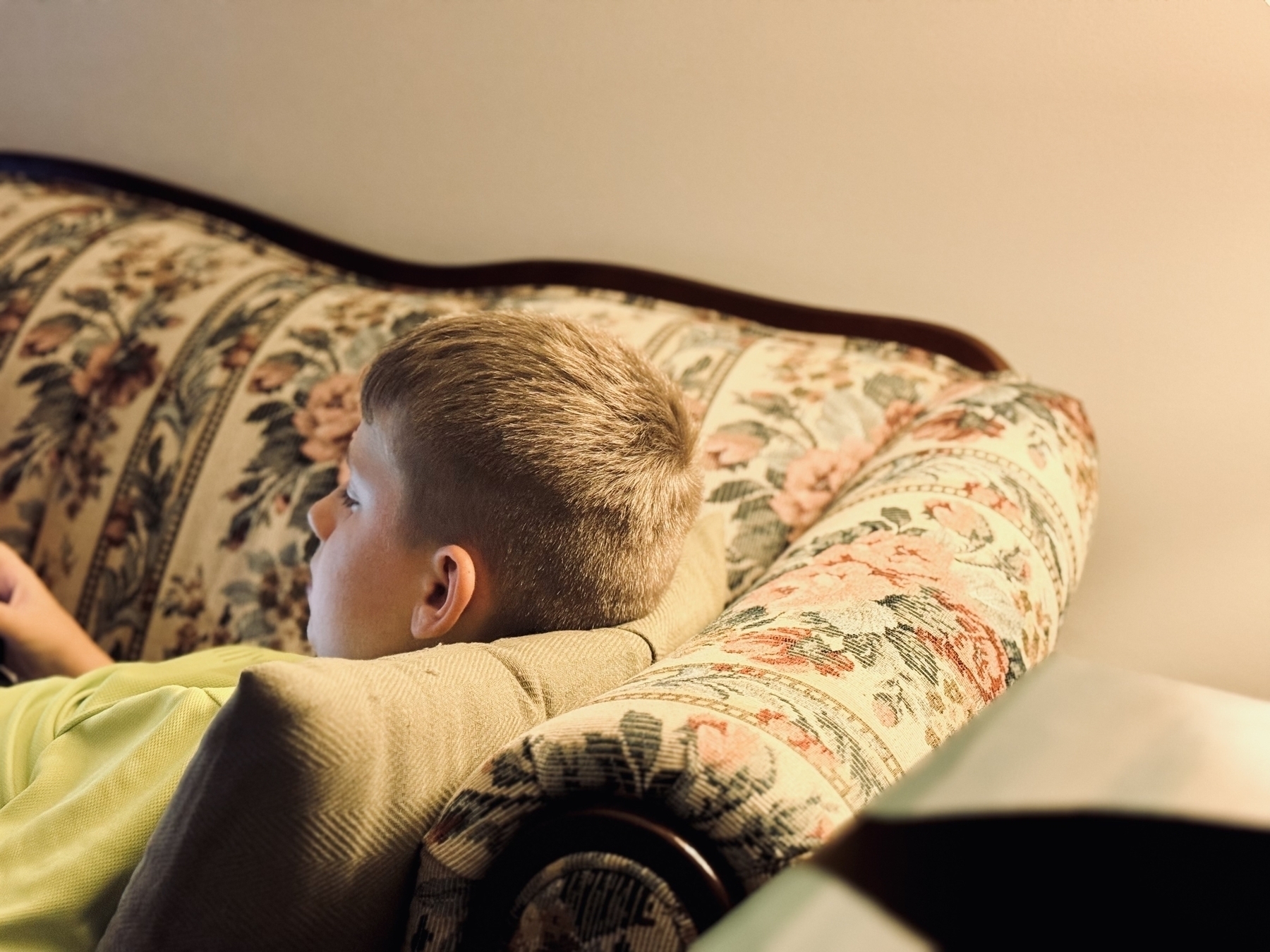 A boy with light hair is lying on a patterned sofa, using an electronic device.