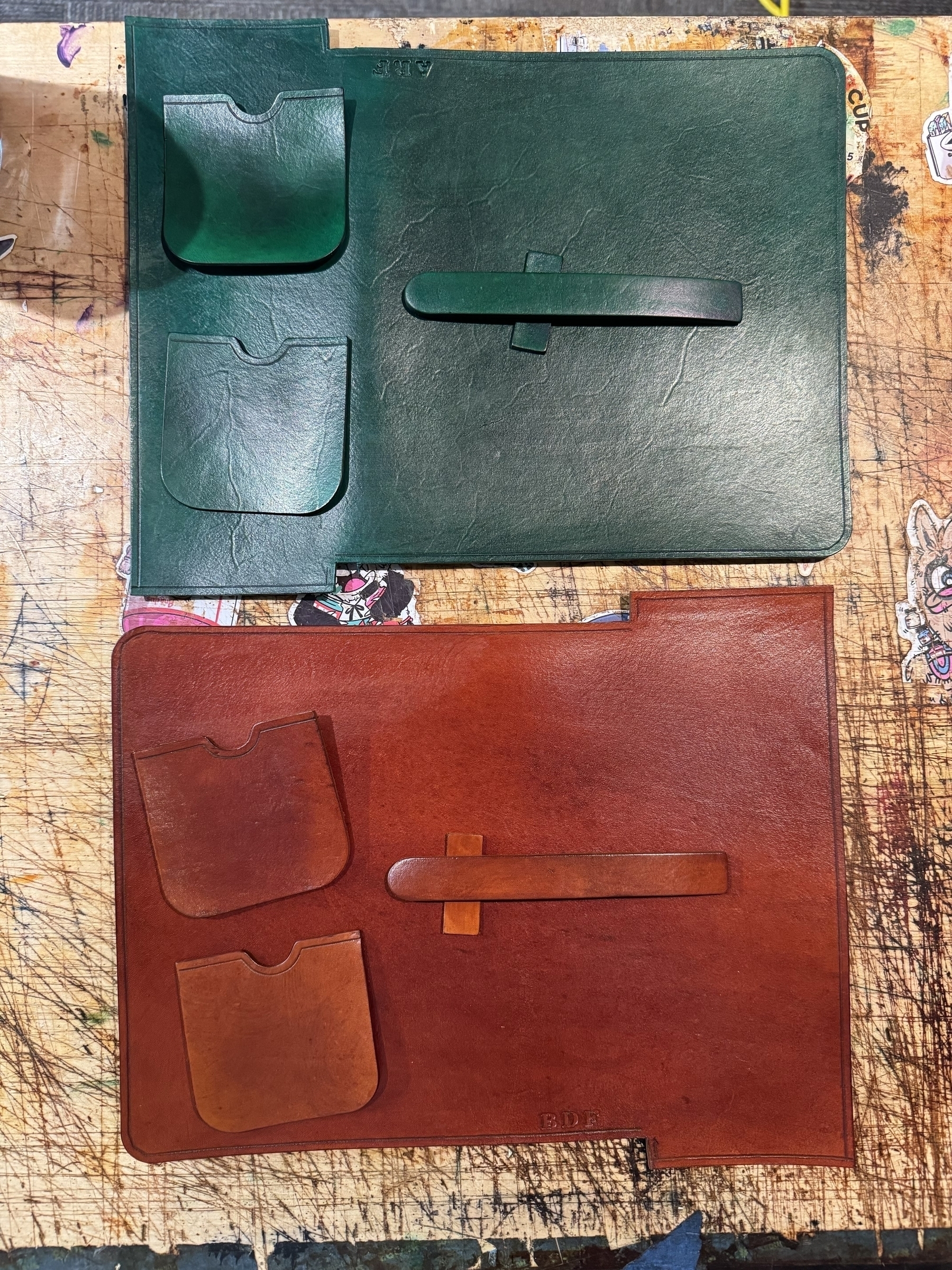 Two leather cigar cases, one green and one brown, are laid out on a wooden surface, each featuring a pair of pockets and a strap.