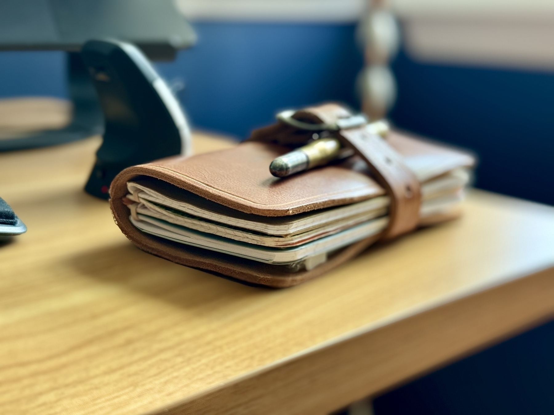 A leather-bound notebook with a pen attached is lying on a wooden desk.