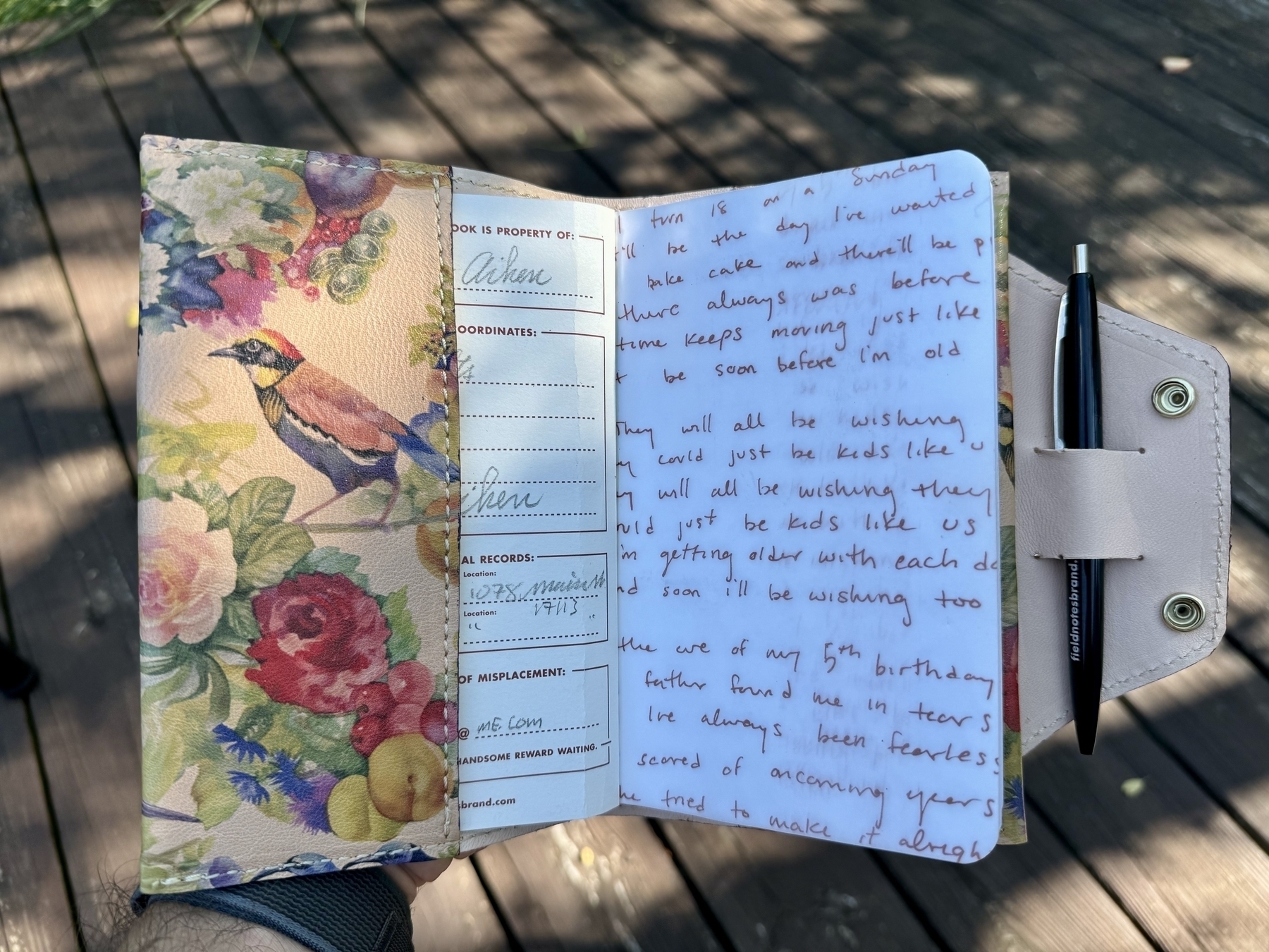 A colorful journal with a bird and floral cover is open to handwritten entries and includes a pen attached to the side.