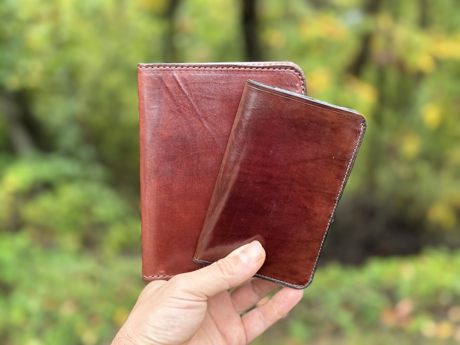 A hand is holding two leather cases of different sizes against a blurred outdoor background.