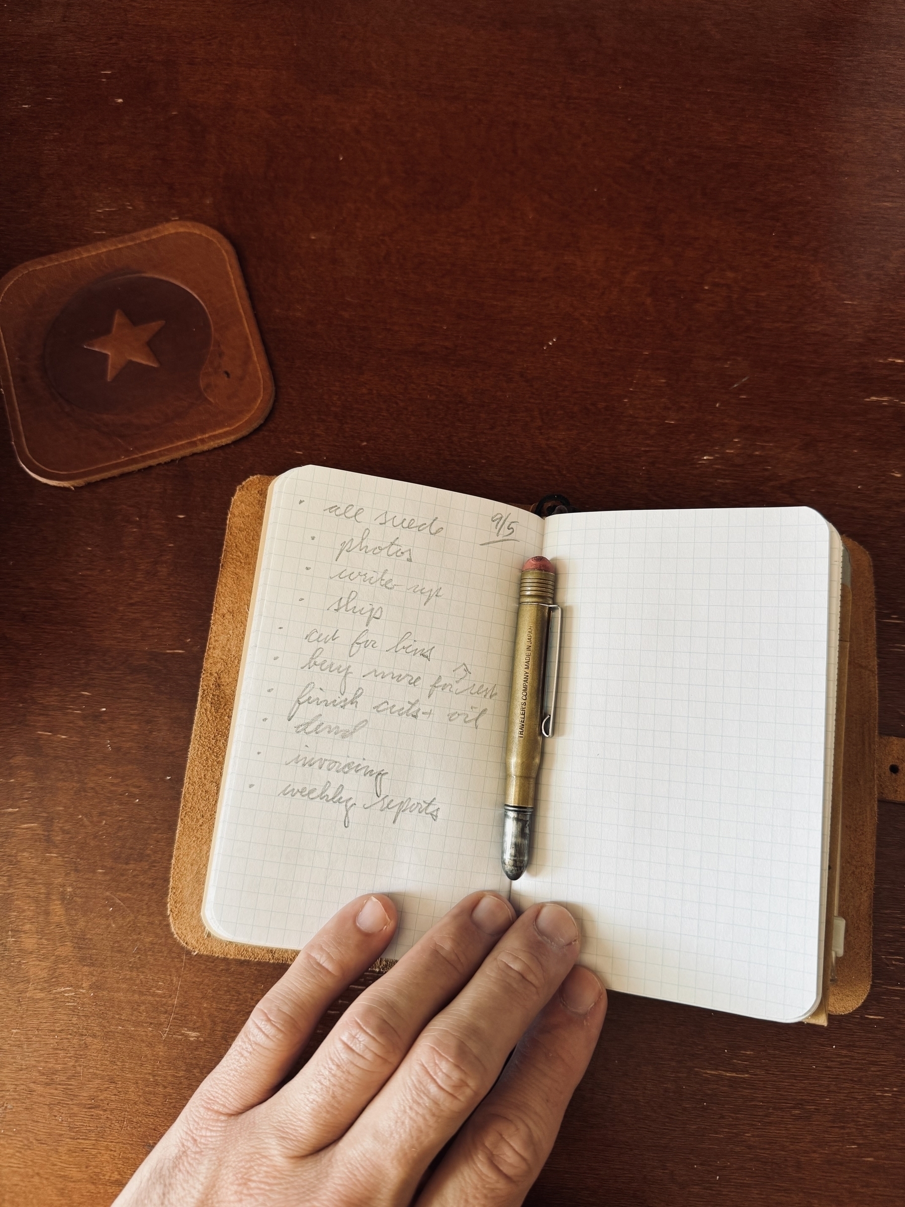 A hand rests on an open notebook with a handwritten list and a pen, placed on a wooden surface next to a square coaster with a star design.