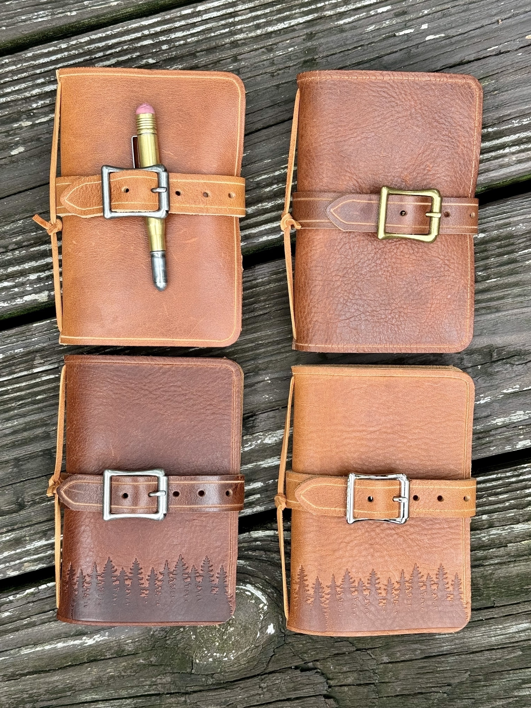 Four leather notebooks with buckled straps, two featuring forest engravings along the bottom, are displayed on a wooden surface.