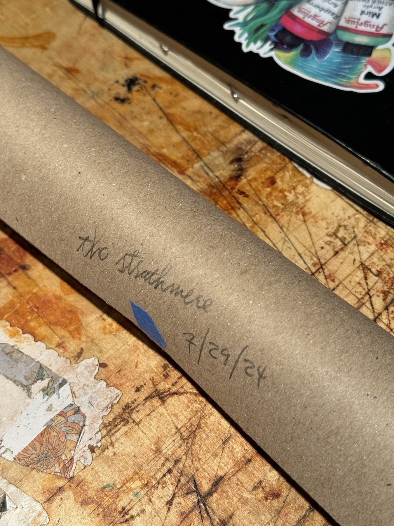 A cardboard tube labeled "The Strathmere 7/29/2024" is resting on a cluttered, paint-stained surface near a piece of art and a sticker-covered item.