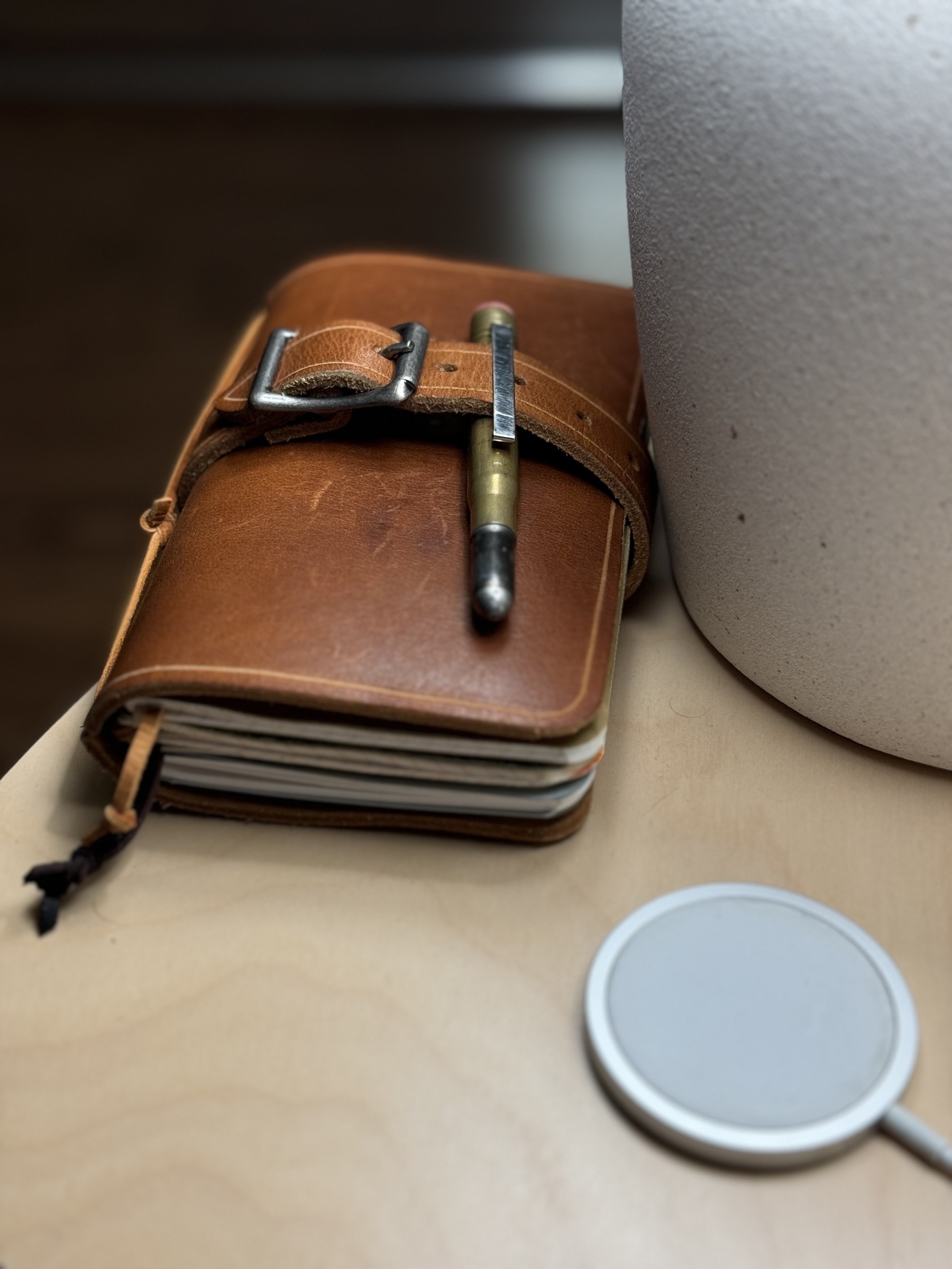 A leather-bound notebook with a pen secured in its strap lies on a wooden surface next to a large white container and a circular gray item.