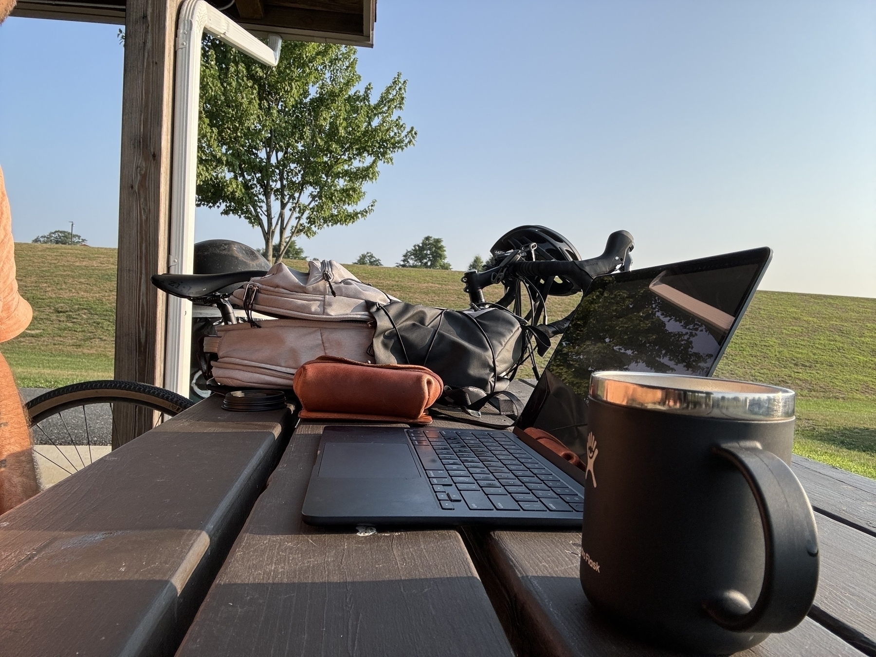 A laptop, a black mug, and a bag are placed on a picnic table with a bicycle parked nearby, set against a backdrop of green grass and clear sky.