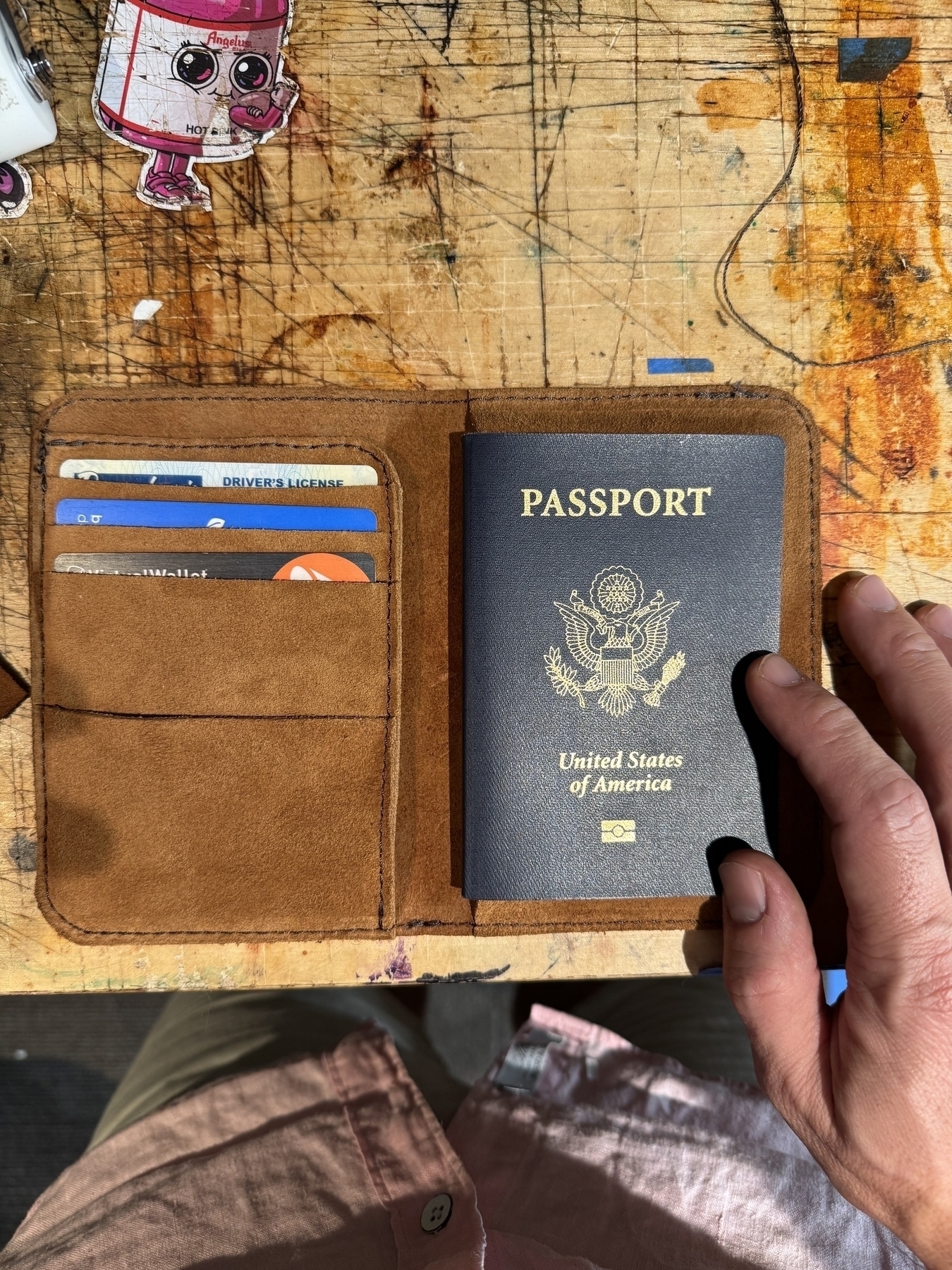 A hand holds a United States passport inside a brown leather case on a wooden desk with several cards visible.