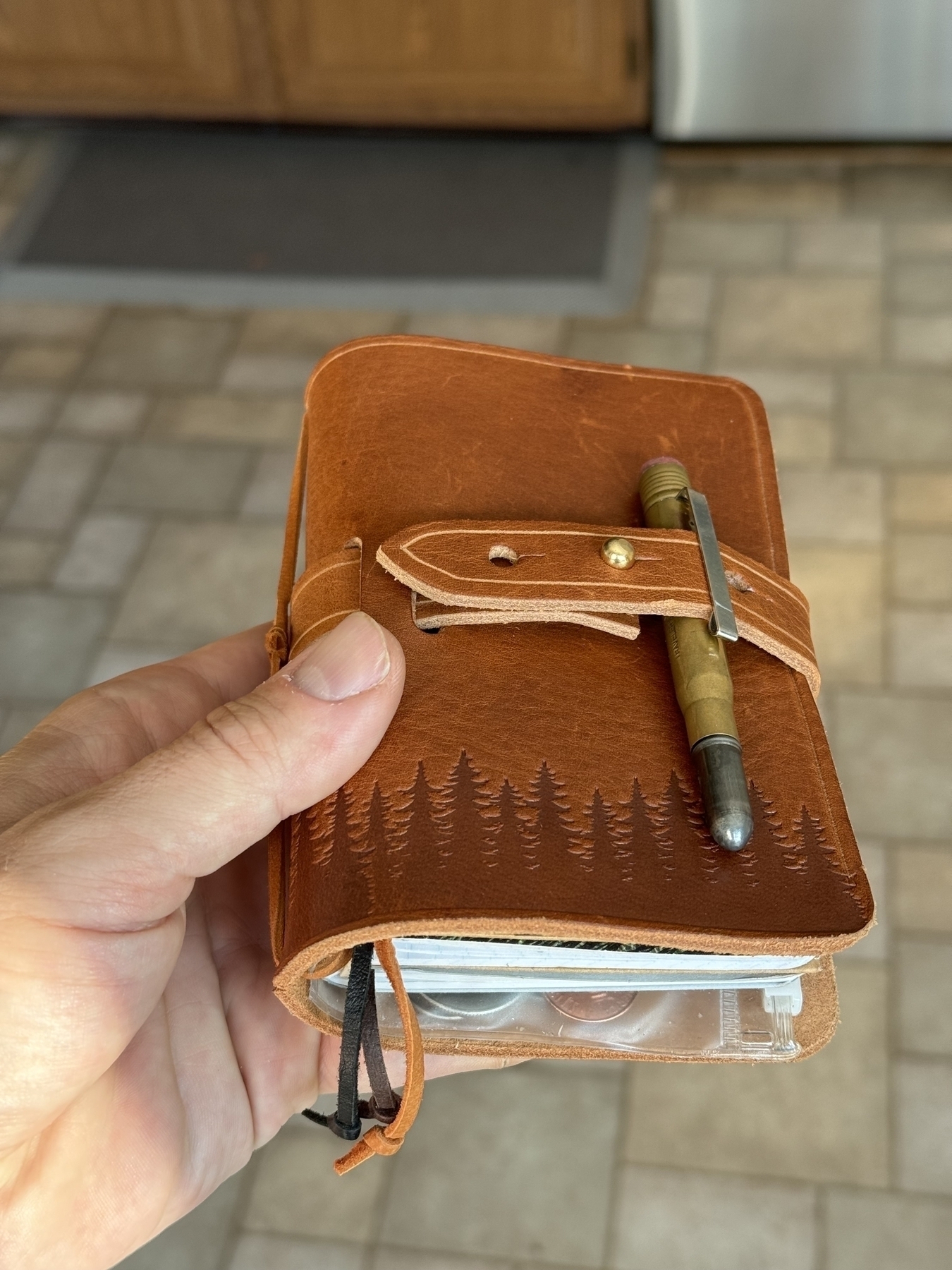 A hand is holding a brown leather wallet with a forest design, secured by a strap, and a pen tucked inside.