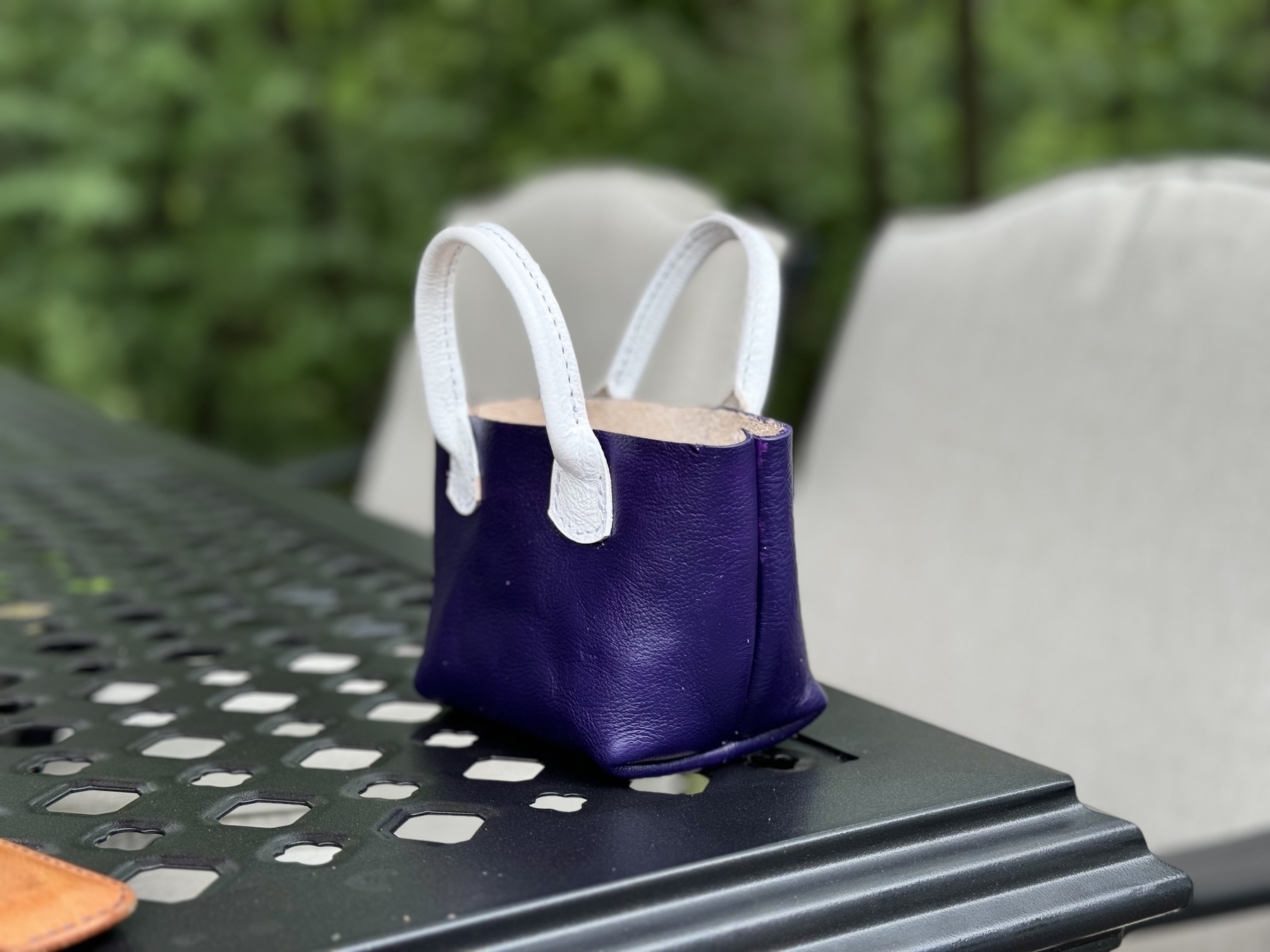 A small purple handbag with white handles is placed on a perforated metal table outdoors, near some chairs and greenery.