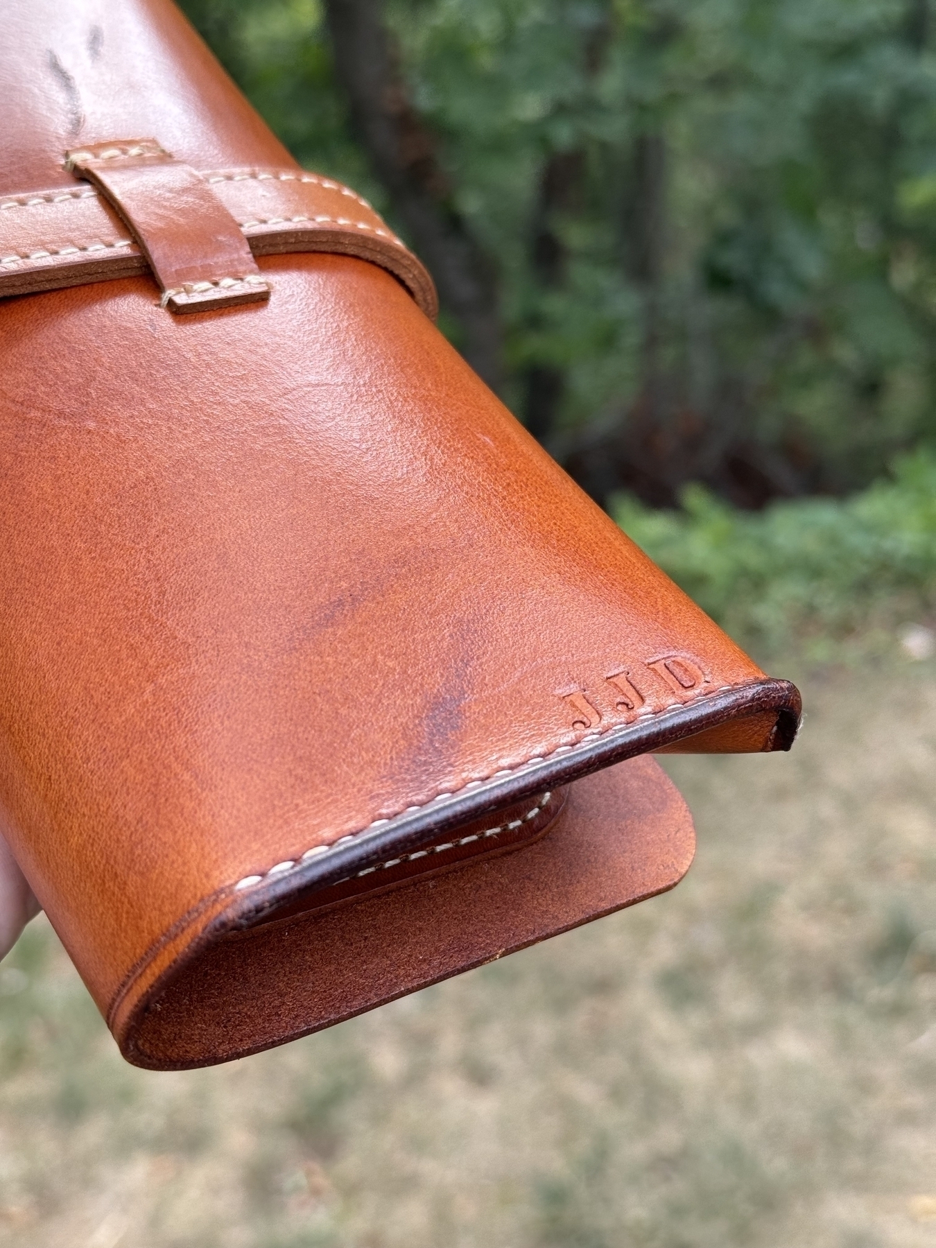 A person is holding a brown leather cigar case with initials "JJD" embossed on one end, blurred foliage in the background.