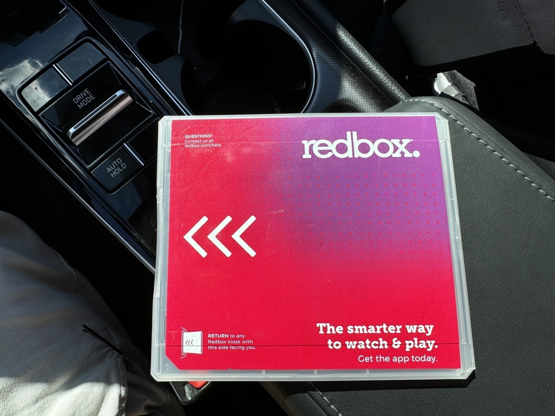 A Redbox DVD case is resting on a car seat, next to some car controls, promoting the convenience of returning it to any Redbox location or dropping it in the slot.