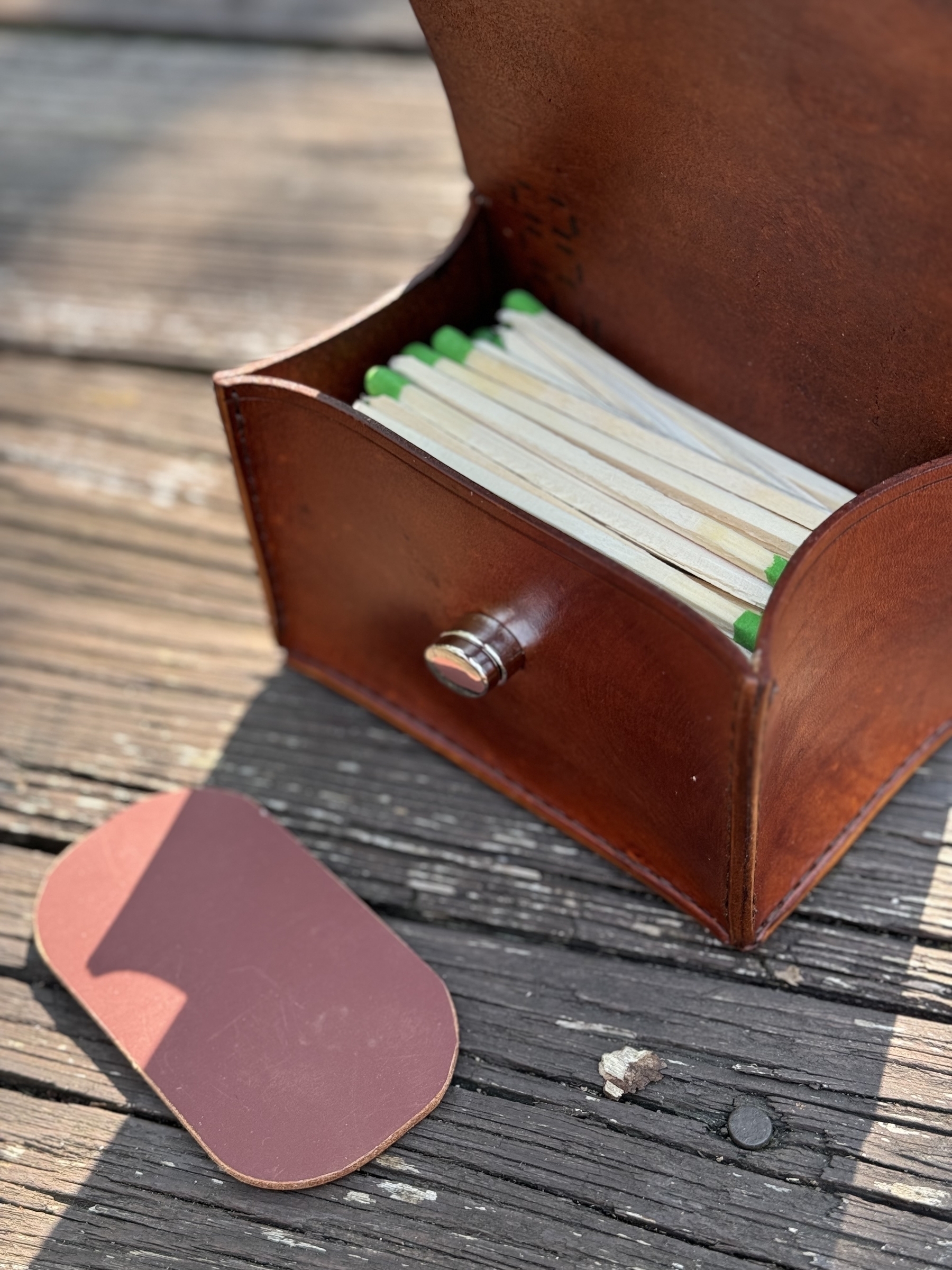 A brown leather case holding green-tipped matches sits on a wooden surface with a matching brown lid resting nearby.