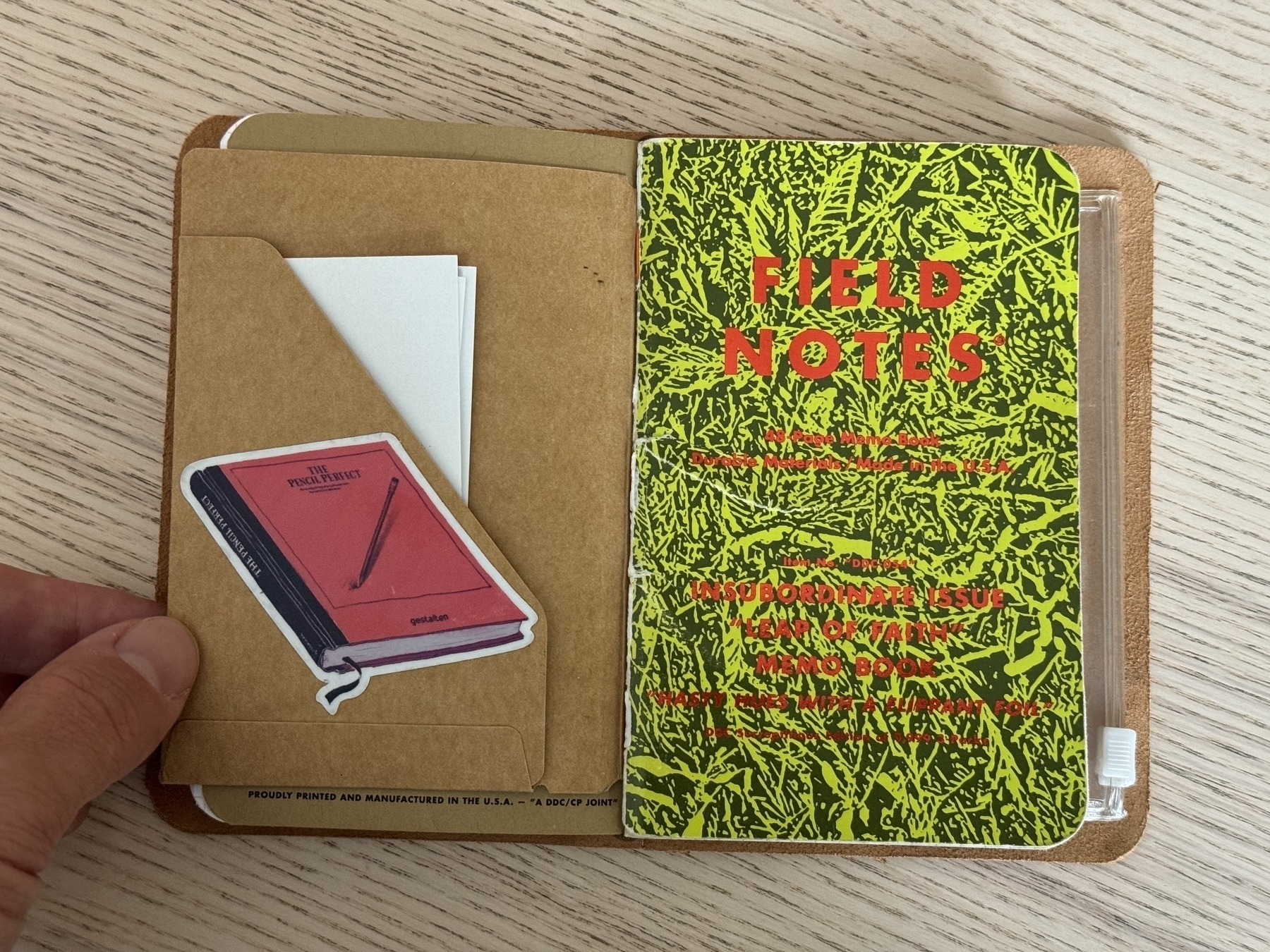 A hand is holding open a notebook cover showing a Field Notes notebook, some paper sheets, and a small sticker of a red journal inside.