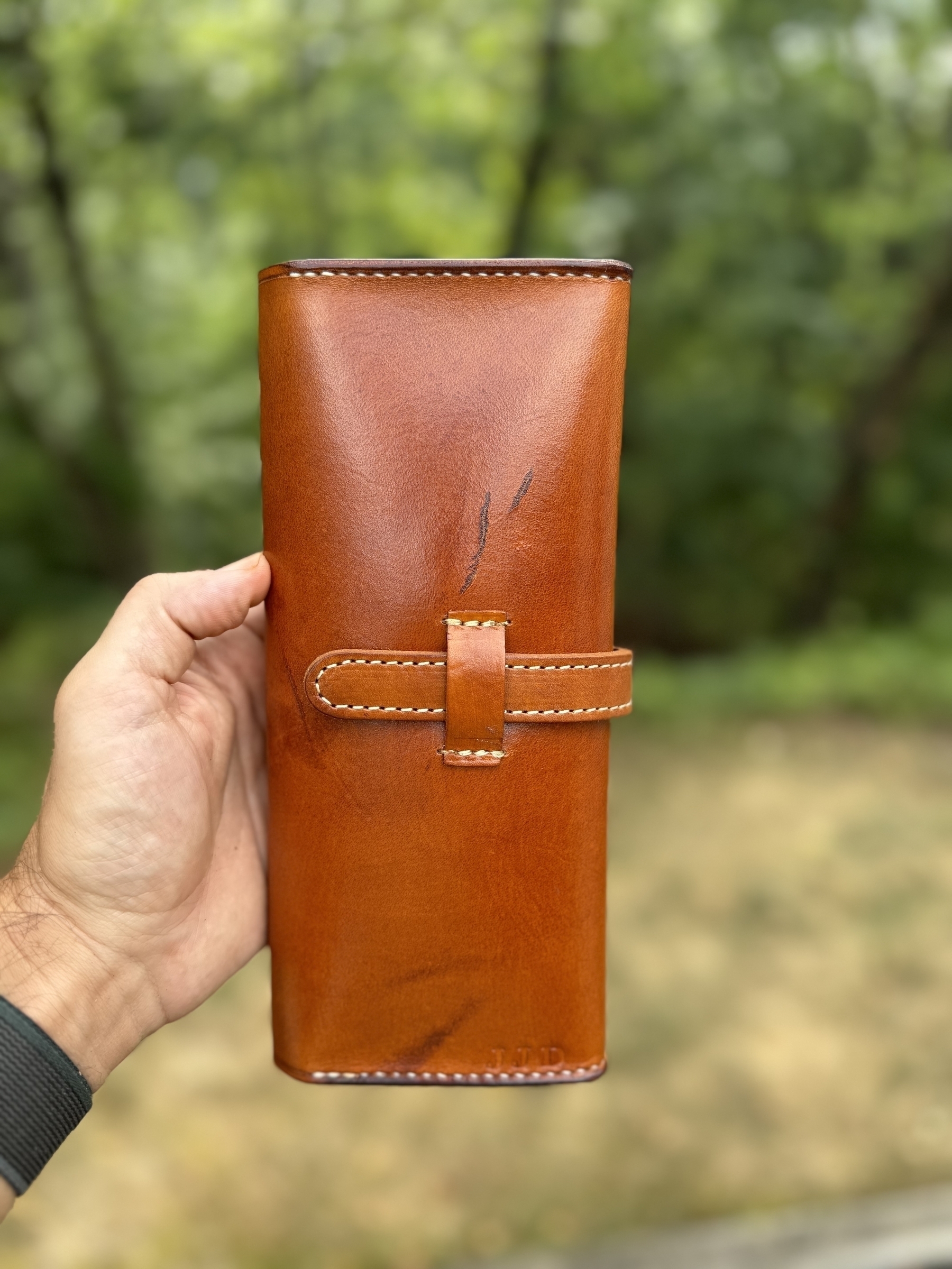 A person is holding a closed, hand-dyed in a light tan shade leather cigar case with visible stitching, against a blurred outdoor background.