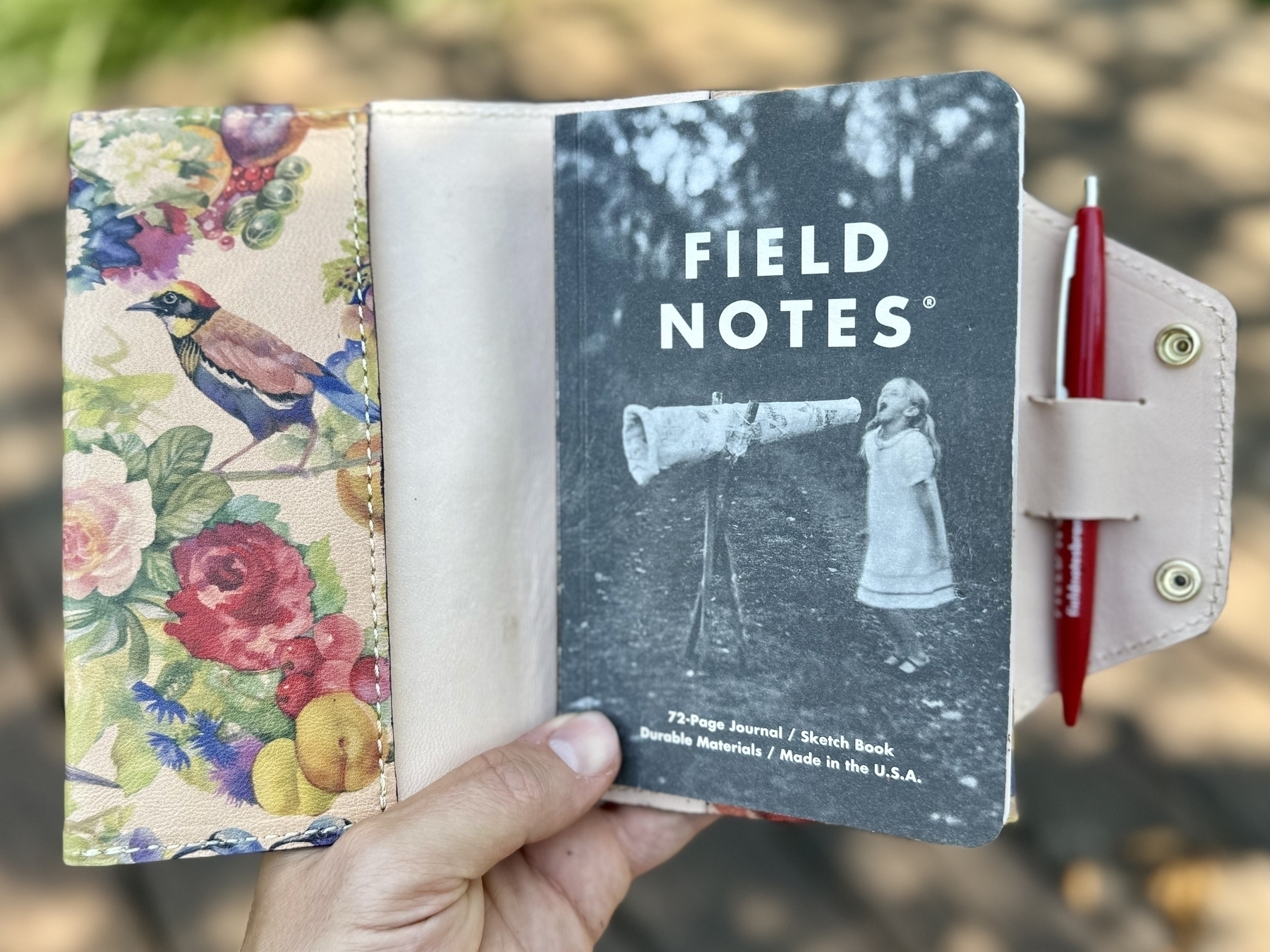 A hand holds an open floral-patterned case containing a small 'Field Notes' notebook and a red pen.