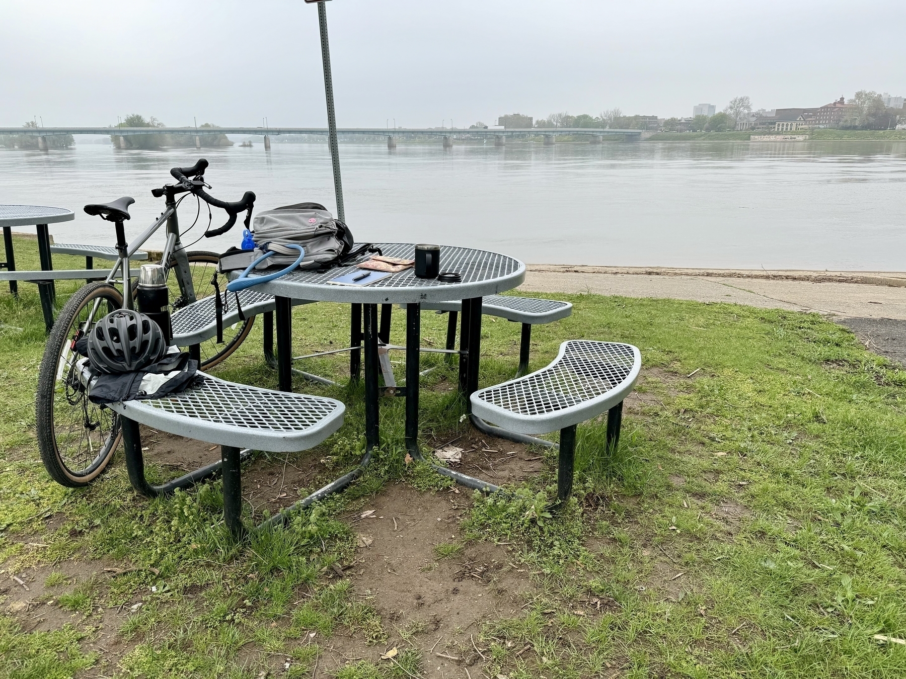Bike leaning up against a picnic table. River in view, and a city on the other side of the river.