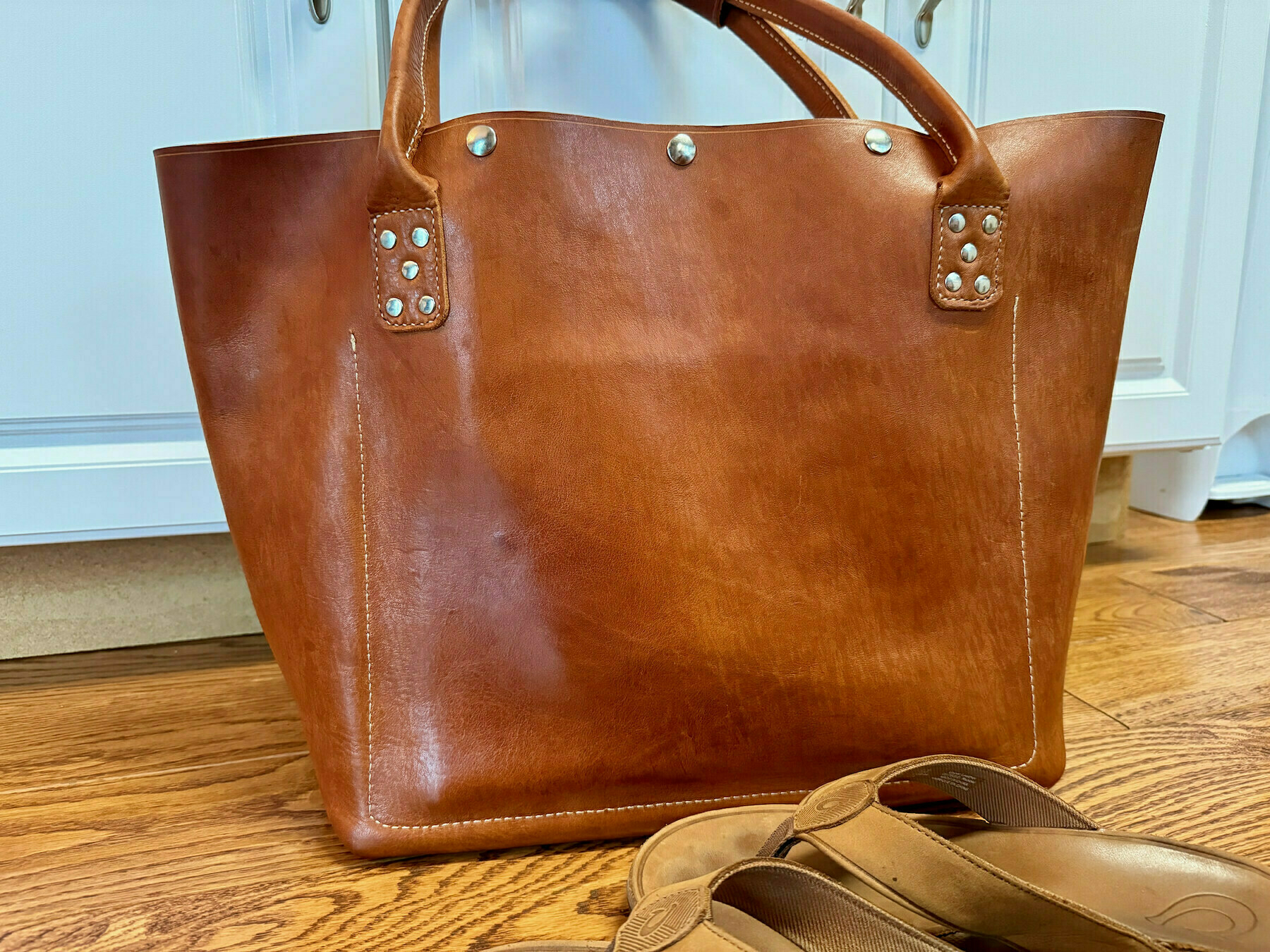A brown leather tote bag with metal accents is placed on a wooden floor beside a pair of matching brown flip-flops.
