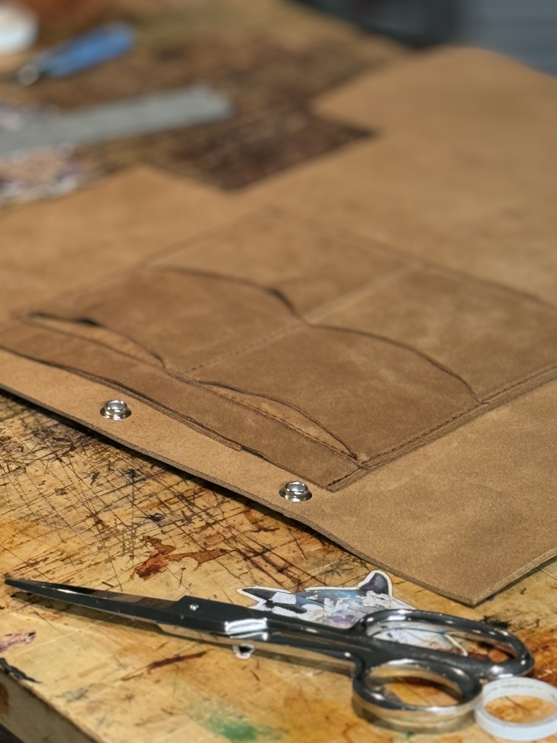 A pair of scissors rests on a scratched wooden workbench next to a piece of tan leather in the process of being crafted.