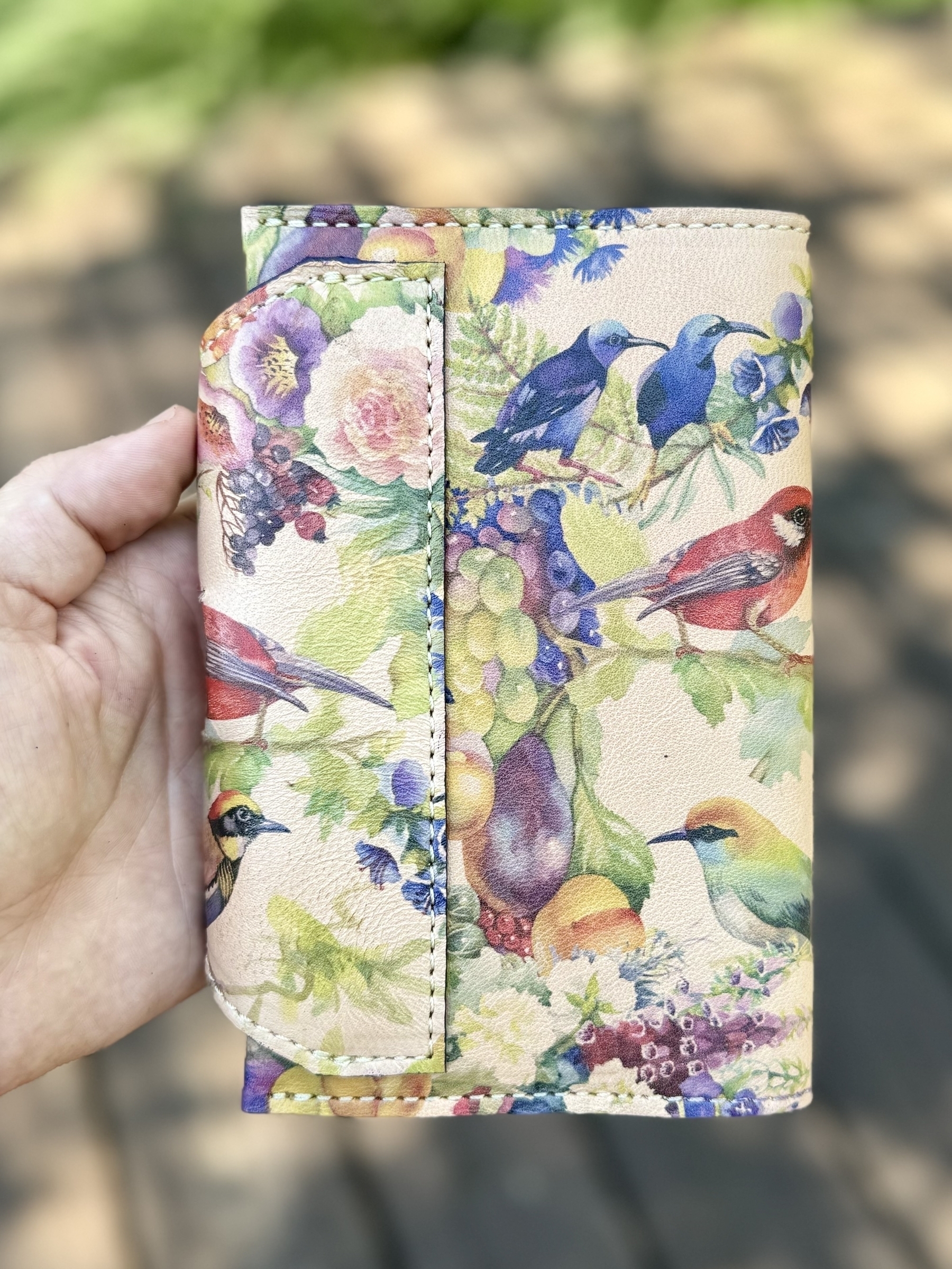 A hand is holding a floral and bird-themed wallet outdoors.