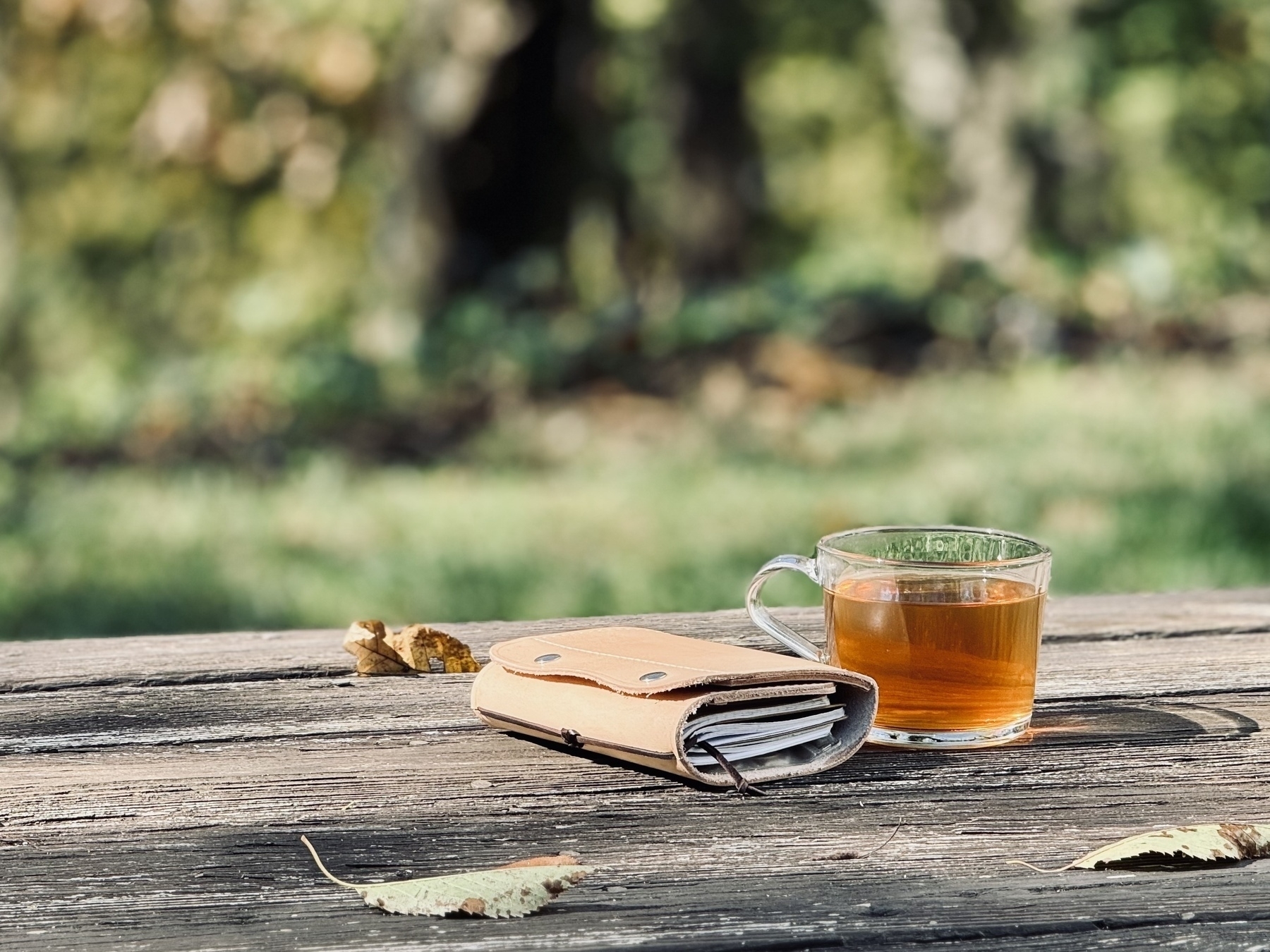 A leather notebook case and a glass of tea rest on a wooden deck outdoors.