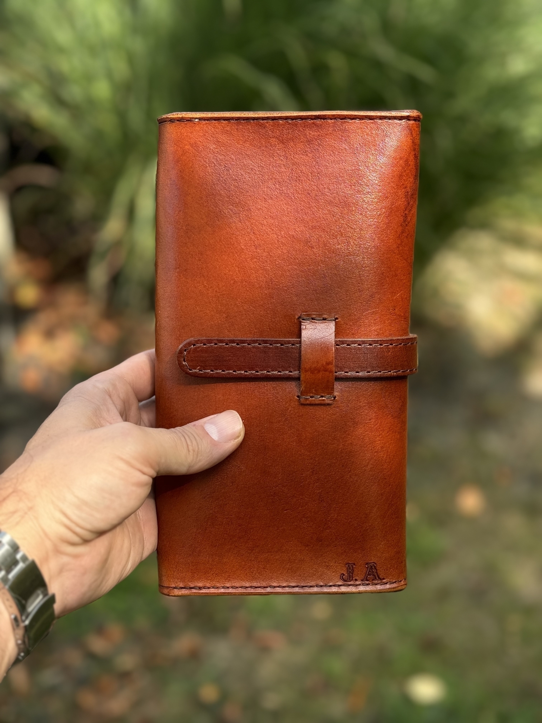 A hand holding a brown leather cigar case with a strap closure, set against a blurred outdoor background.