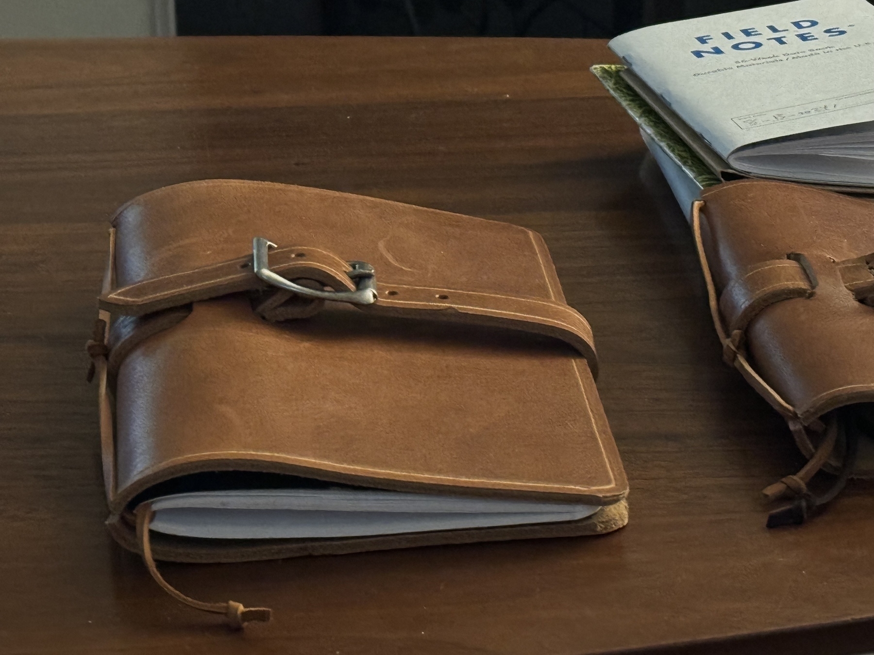 A leather-bound notebook with a buckle closure lies on a wooden table next to a smaller leather notebook and a "Field Notes" notepad.