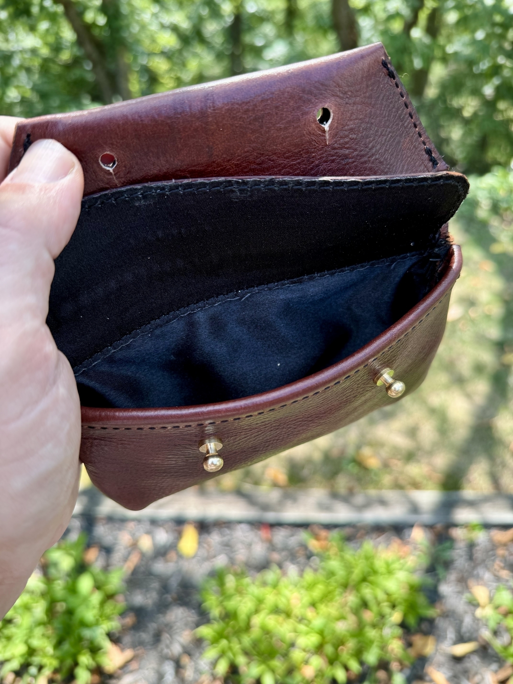 A person is holding an open, small, brown leather pouch with a black interior lining in an outdoor setting.