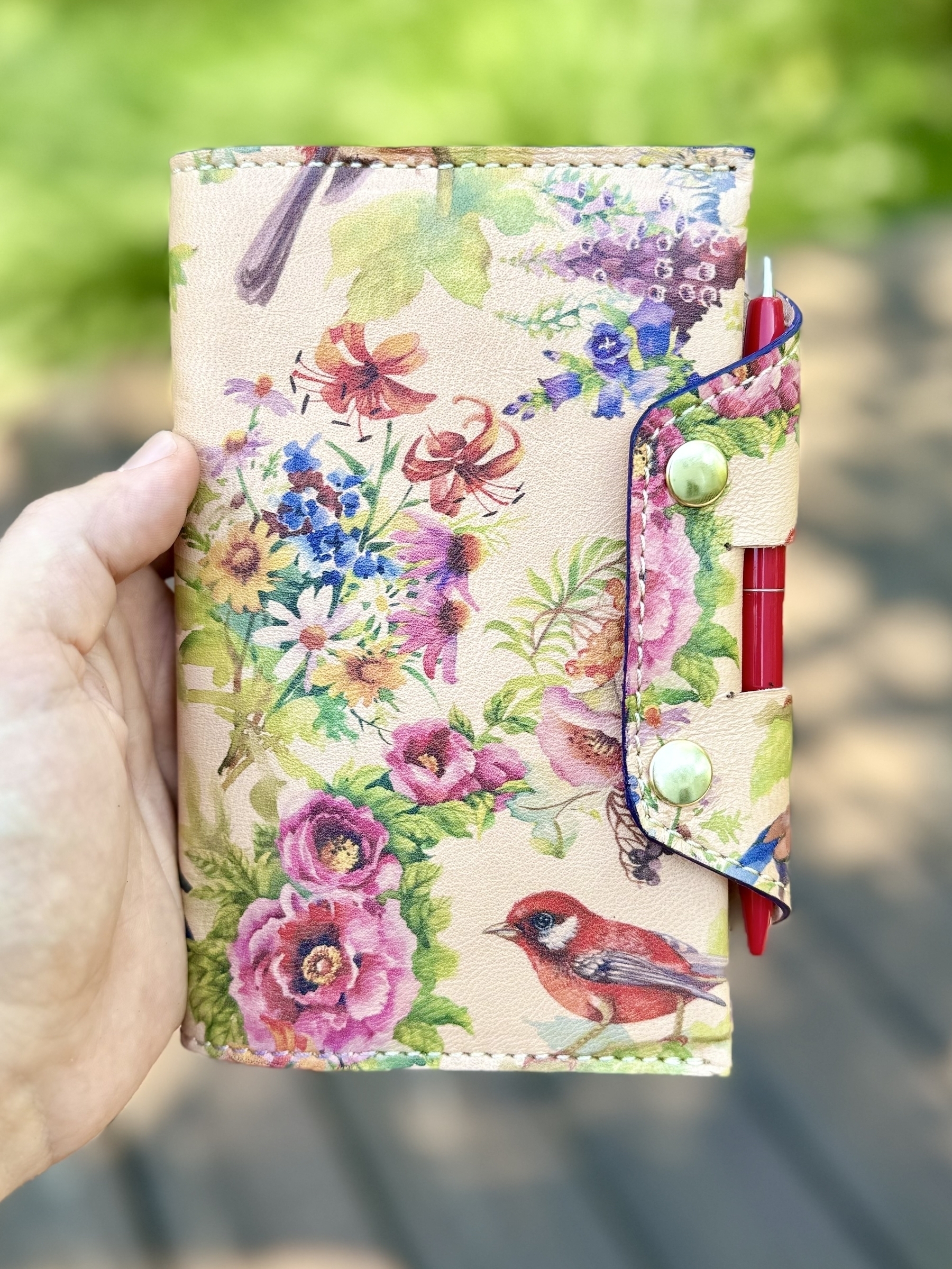 A hand is holding a floral-patterned notebook with a red pen attached to it.