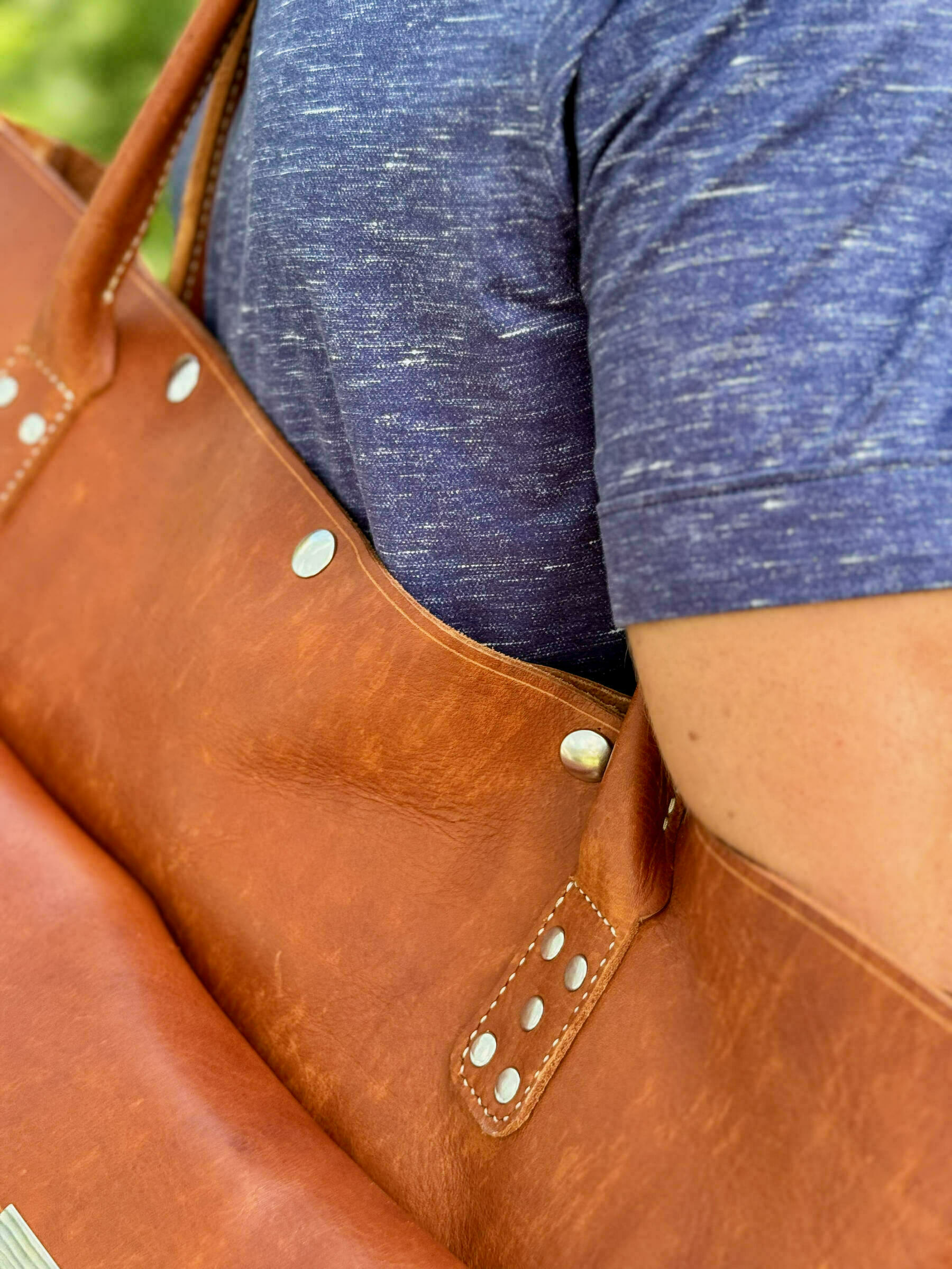 A person wearing a blue shirt carries a brown leather bag with metallic rivets on their shoulder.