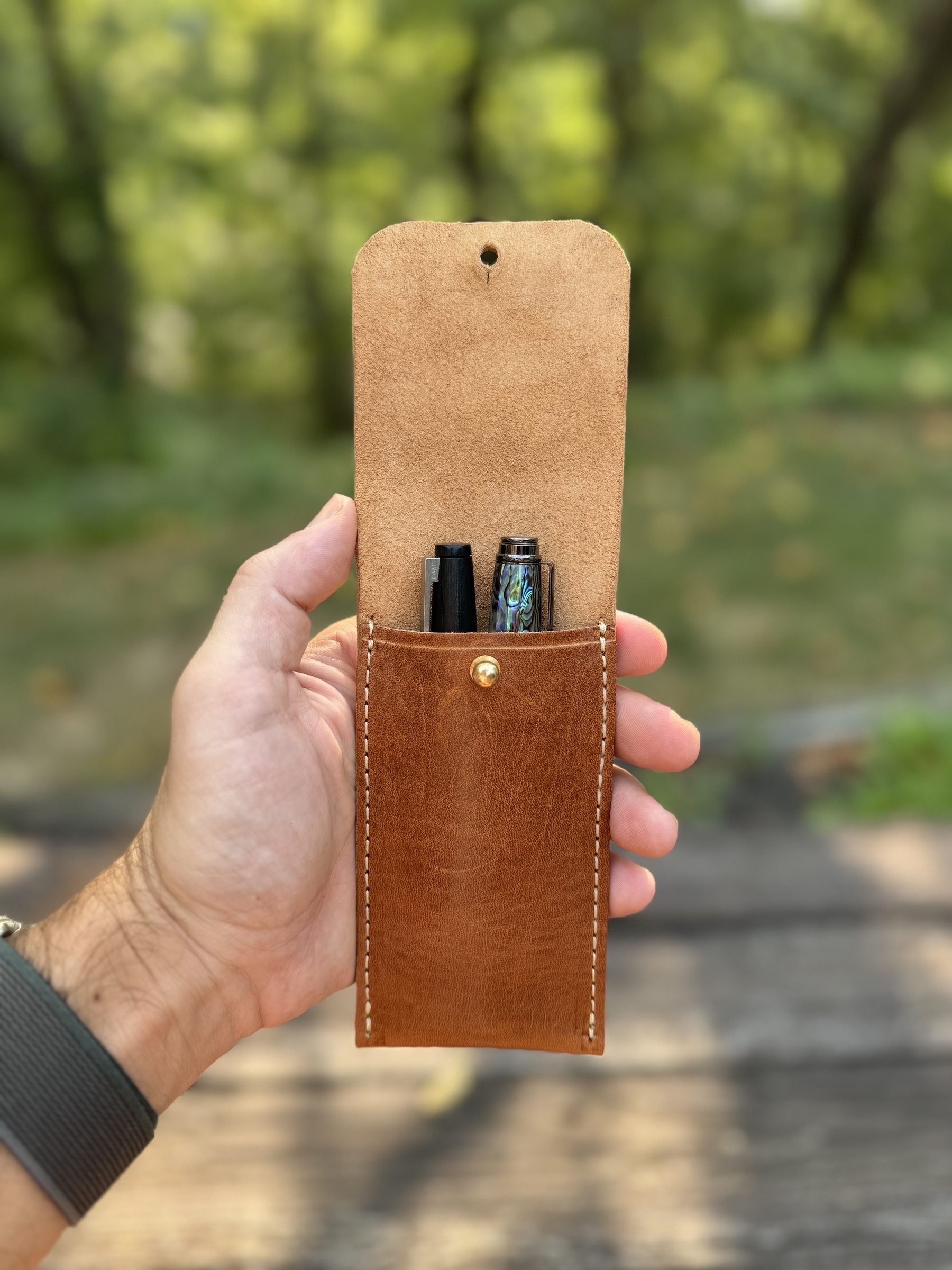 A hand is holding an open leather pouch containing two pens, with a blurred outdoor background.