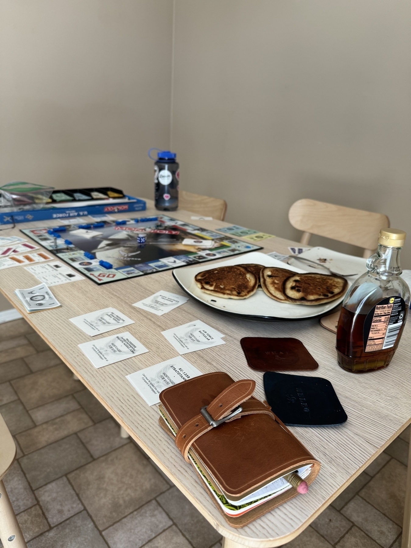 A table is set with a game board, some folders and notes, a water bottle, and a plate of pancakes with syrup.