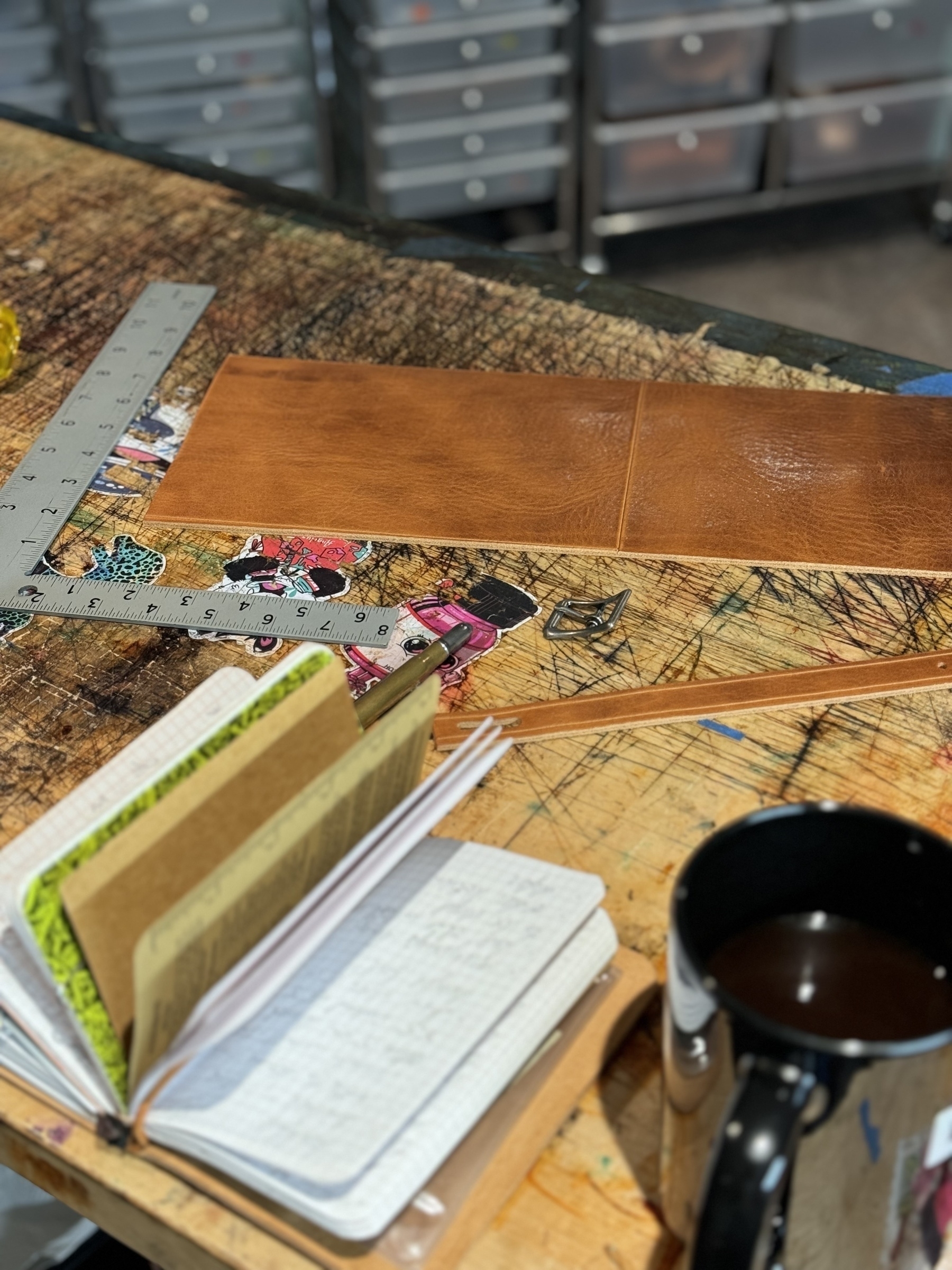 On a cluttered workspace, there are notebooks, a leather piece with rulers, and a cup of coffee.